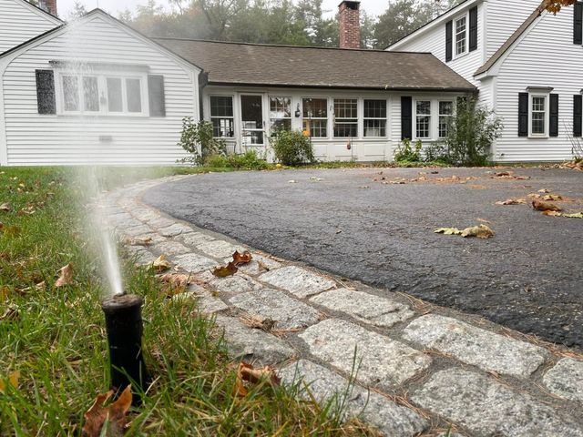 Sprinkler watering a gravel driveway beside a white house with a stone border.