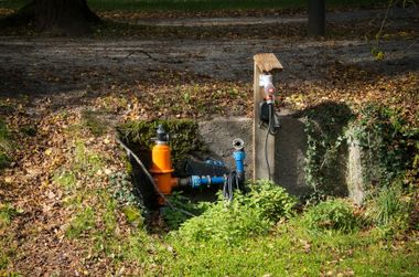 Small outdoor utility pit with pipes, a blue valve, and an orange pump in a grassy ditch.