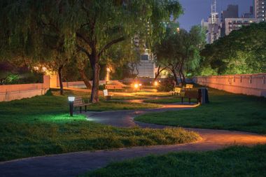Nighttime park path with glowing streetlights, trees, and city buildings in the background
