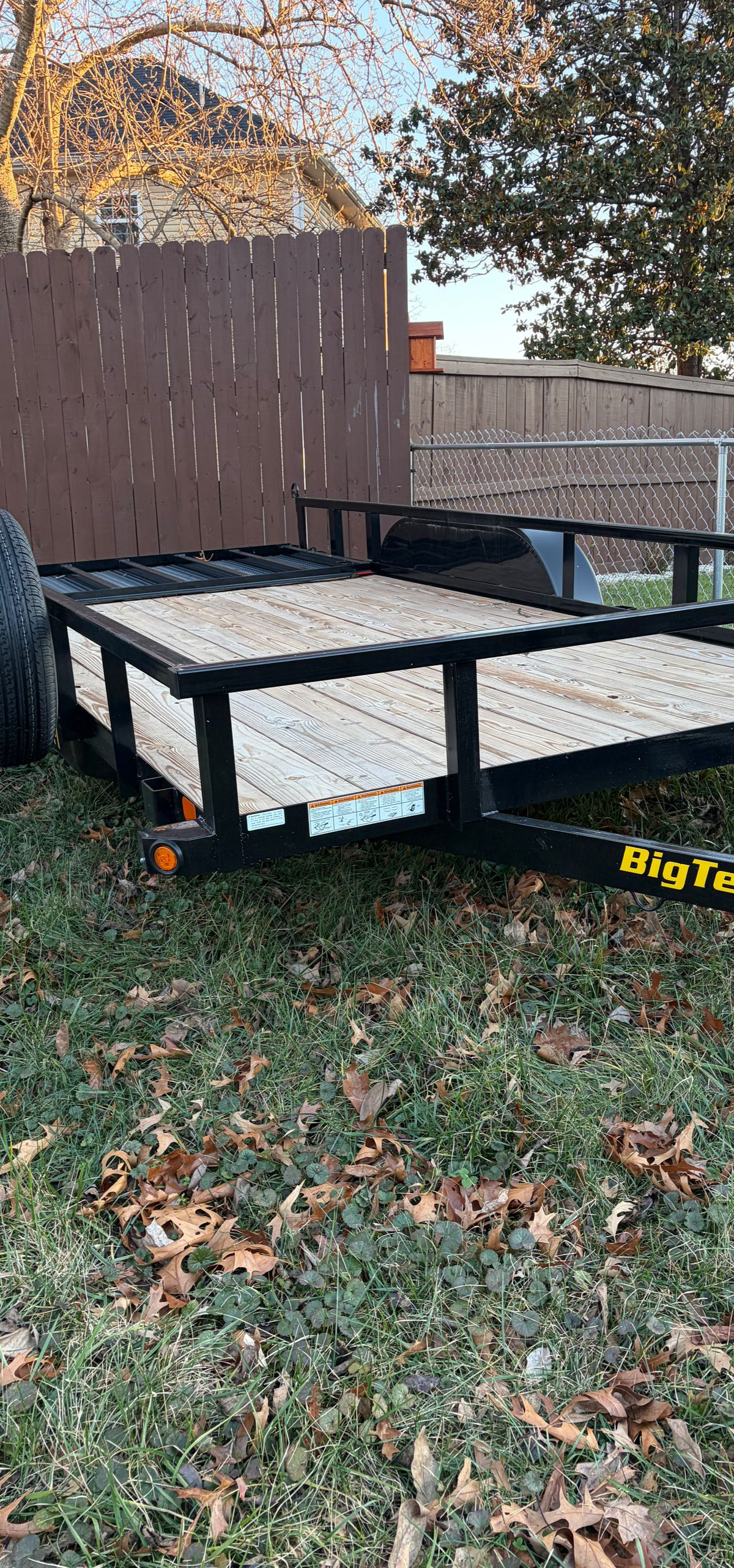 Black utility trailer with white sides parked on grass beside a wooden fence and tree in a yard