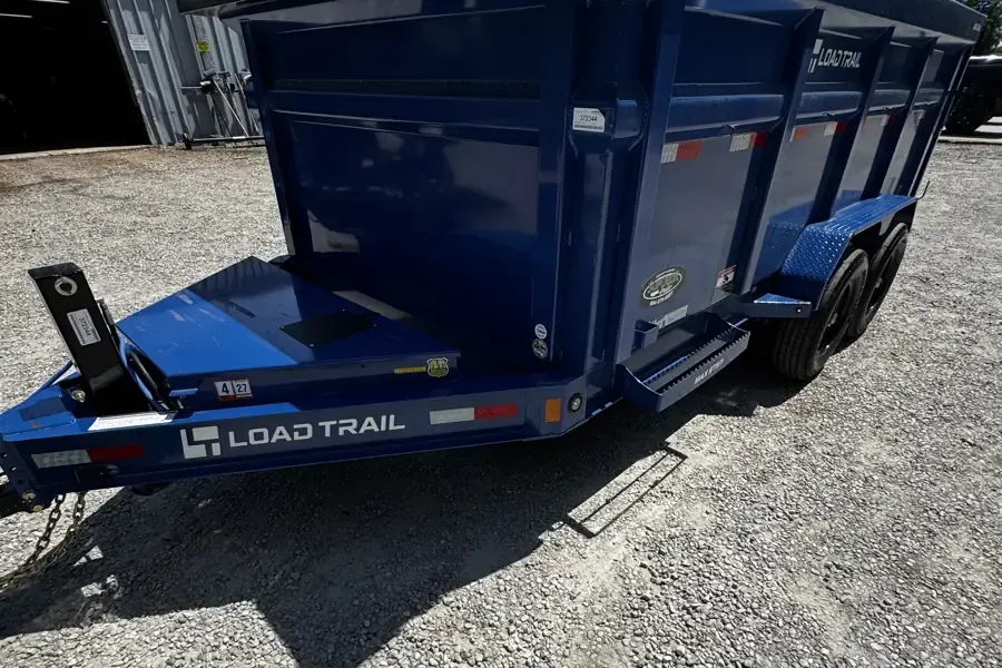 Blue Load Trail dump trailer parked on gravel, side view with black wheels and hitch visible