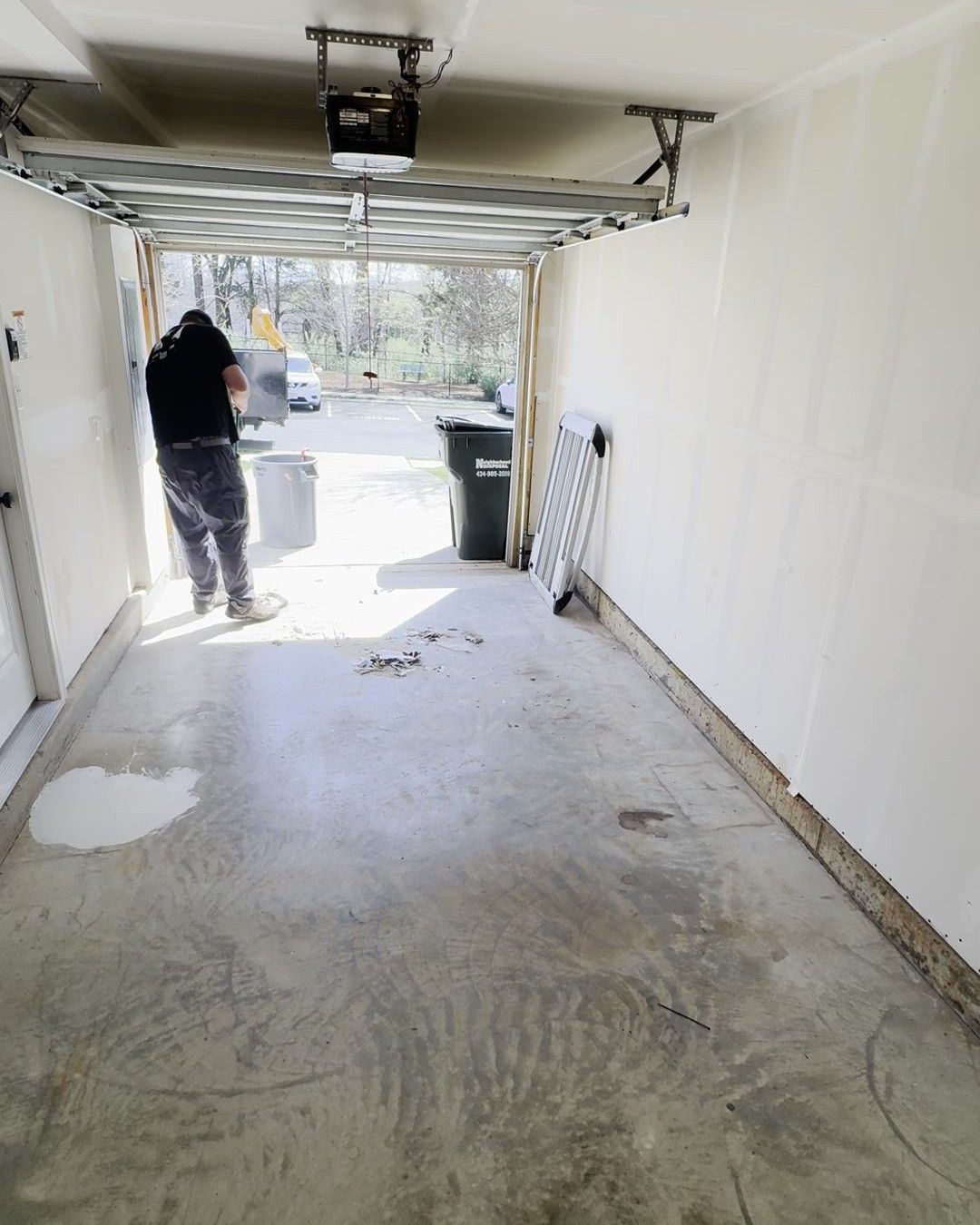 Person in a garage under repair, with bare concrete floor, open door, and tools leaning against the wall.