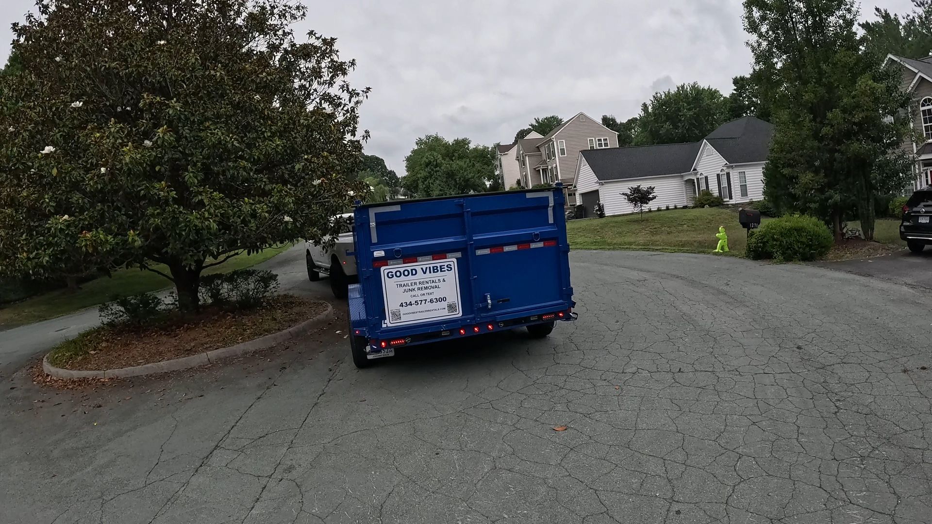 Blue dumpster on a quiet residential street, with houses and trees in the background.