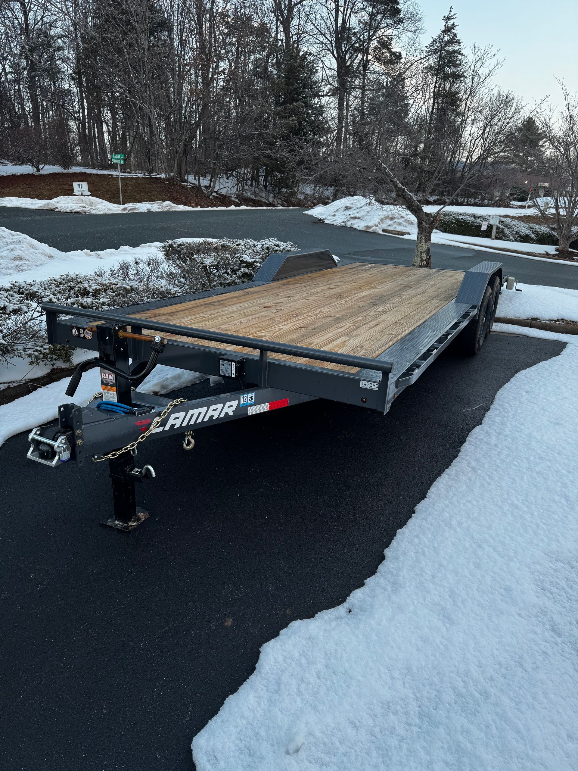 Flatbed utility trailer parked on a snowy driveway beside a blacktop road with snowbanks and trees.