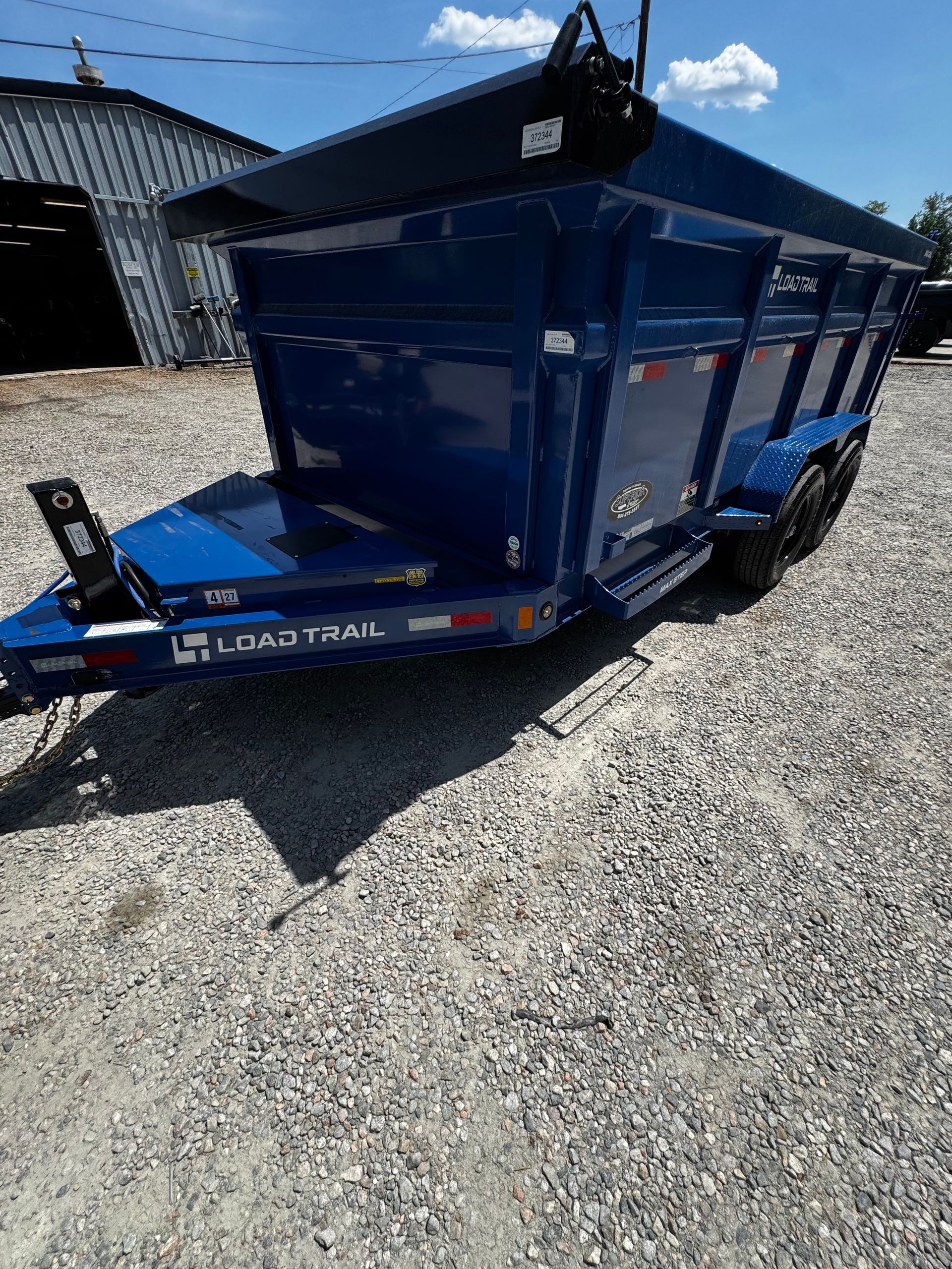 Blue dump trailer parked on gravel beside a building under a sunny sky