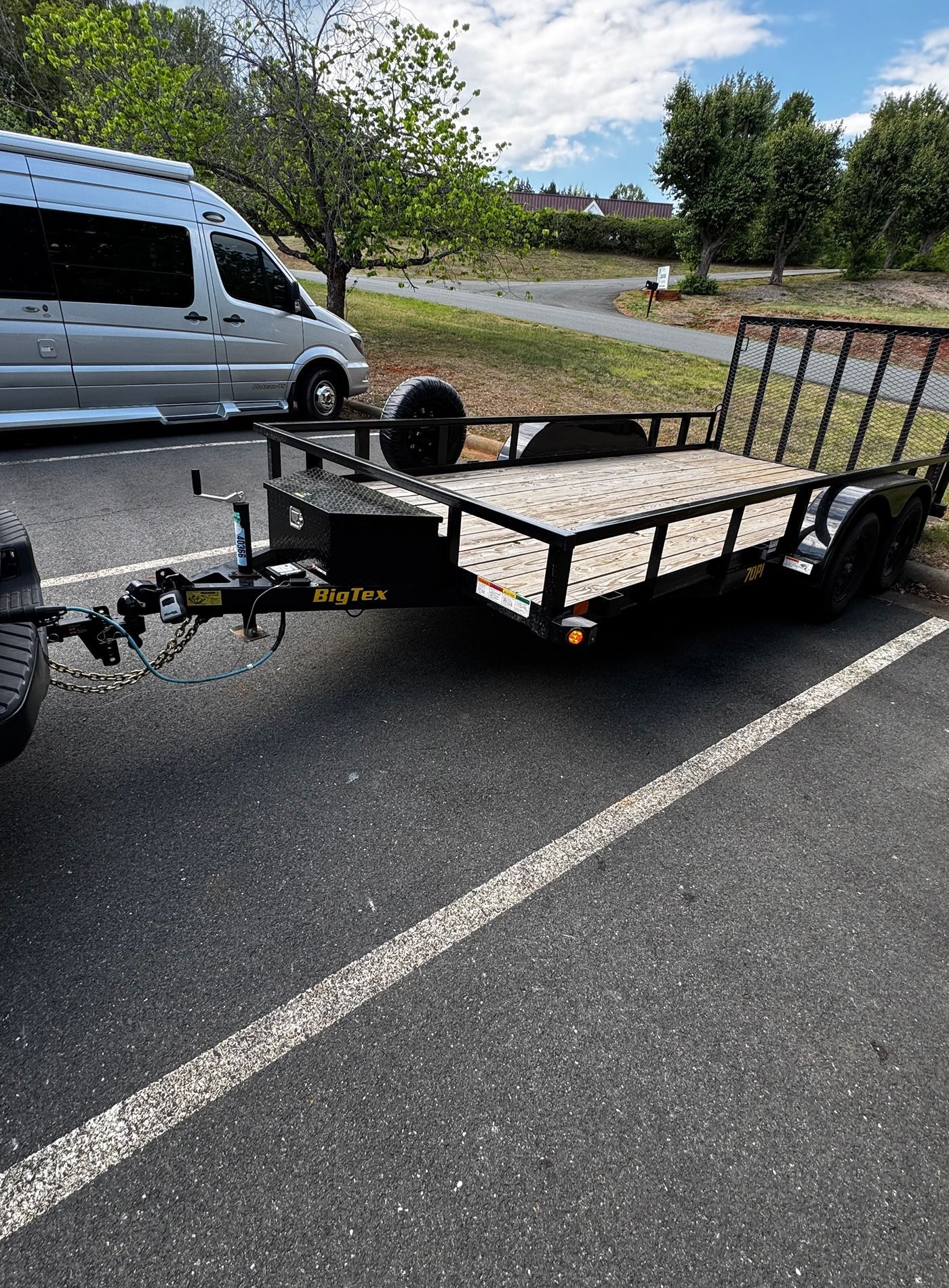 Black utility trailer parked on a road shoulder beside a silver van, with metal rail sides and a textured deck