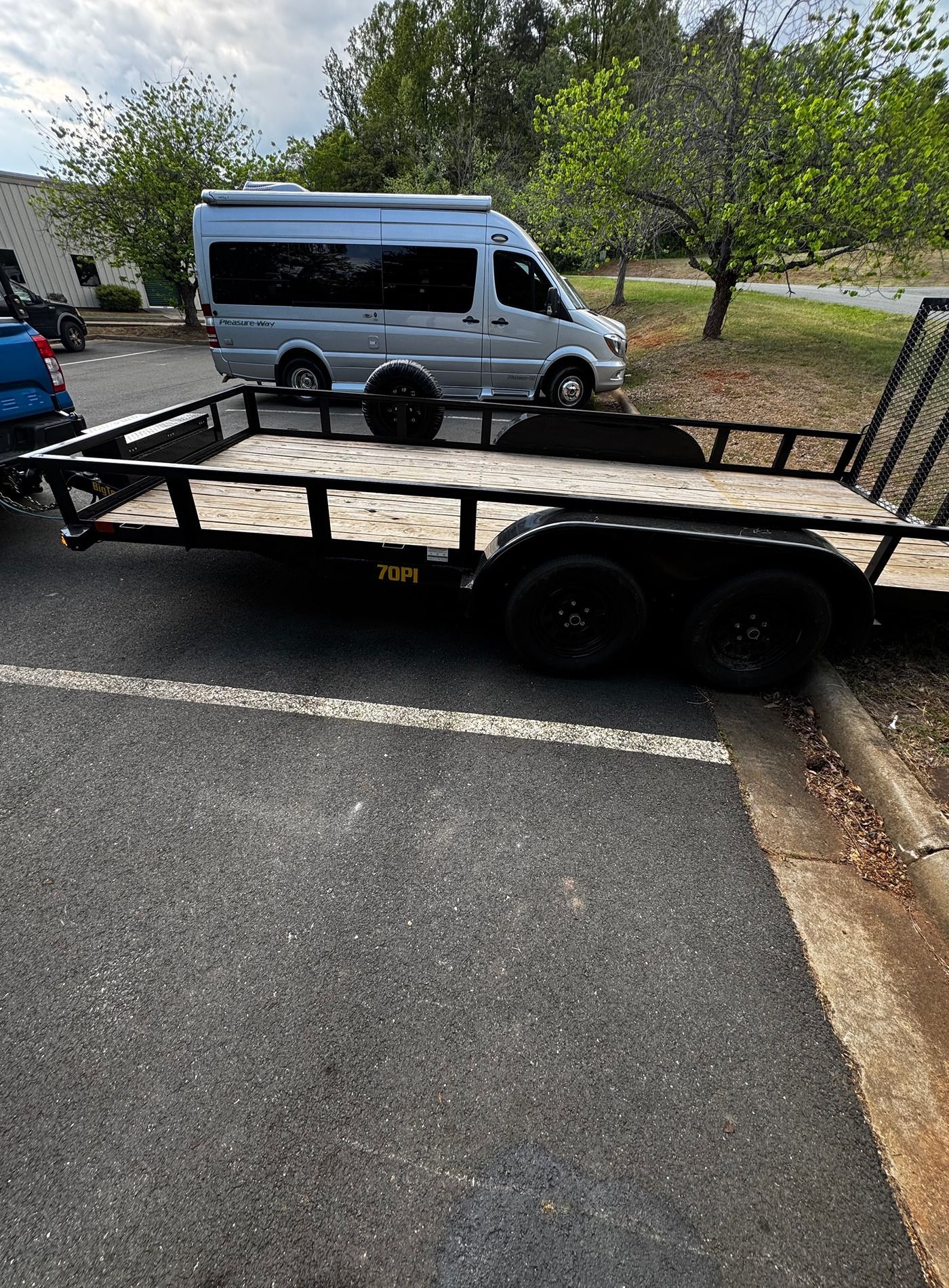 Black utility trailer parked beside a white van on a roadside, with trees and a building in the background