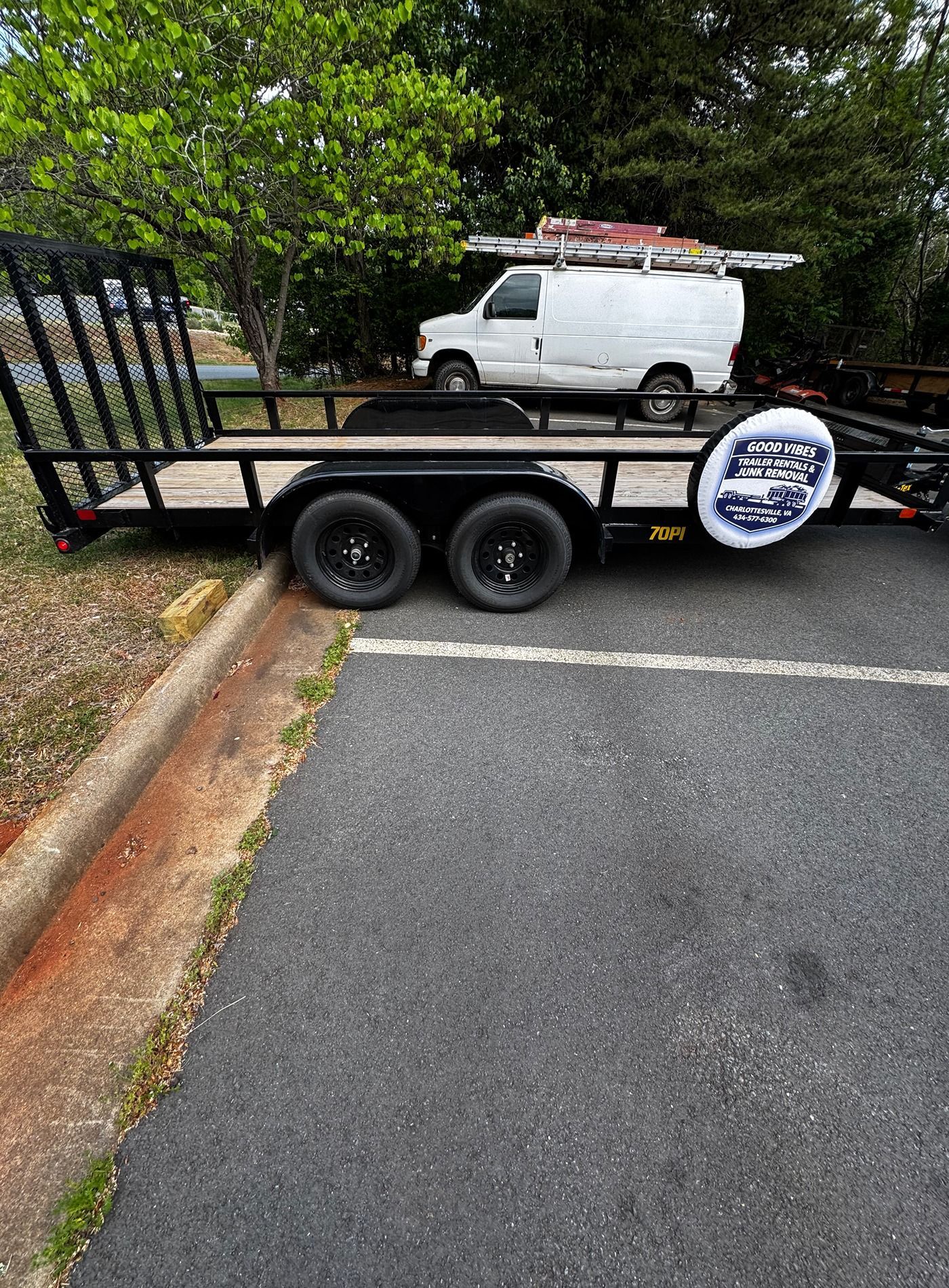 Black utility trailer with mesh ramp parked beside a curb, with a white van and trees in the background.