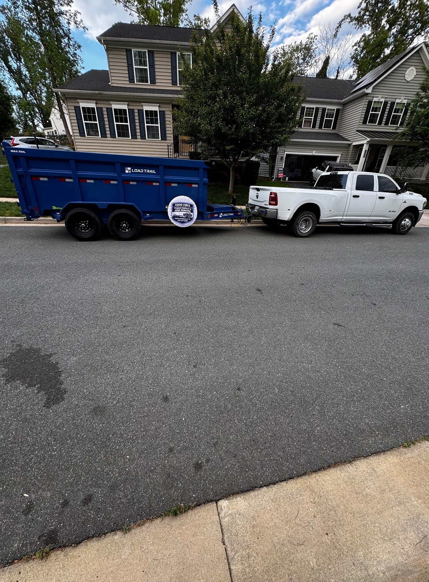 White pickup truck towing a blue dump trailer parked on a residential street in front of houses.
