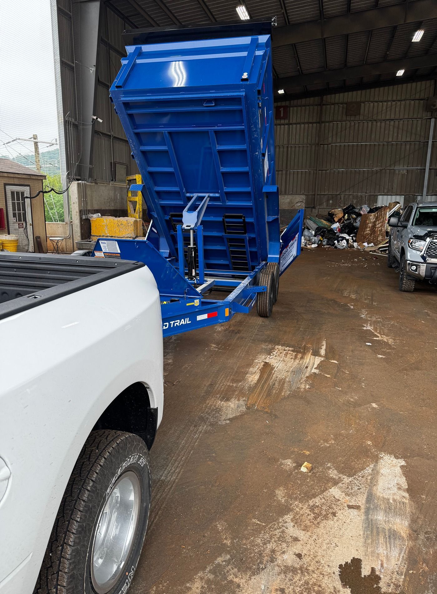 Blue dump trailer in a warehouse, tipped up for unloading beside a white pickup truck.