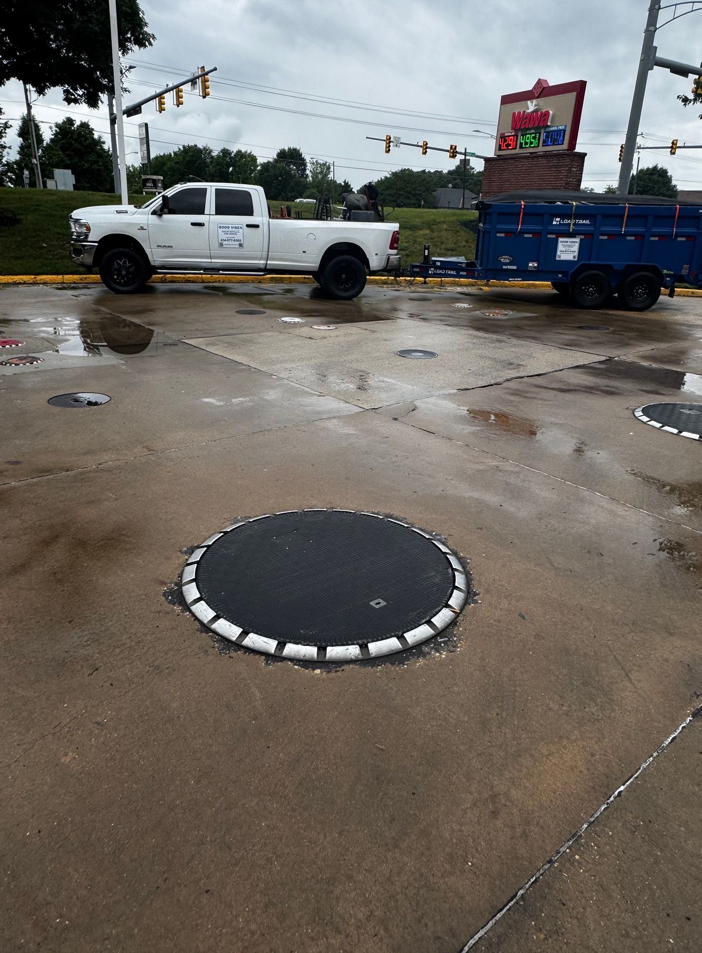 Wet street with a white pickup truck, blue trailer, and round manhole cover in the foreground