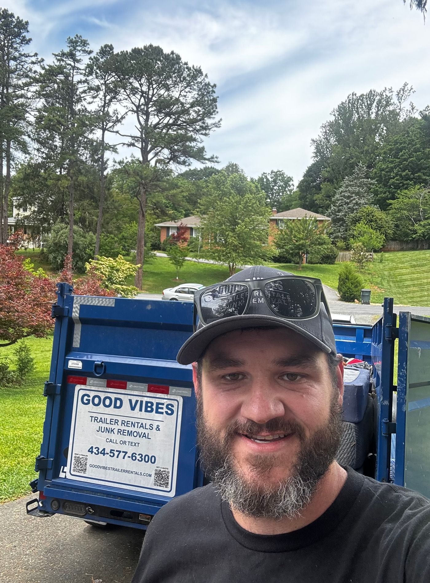 Man smiling in front of a blue dumpster in a leafy yard with a “Good Vibes” sign, carrying debris.