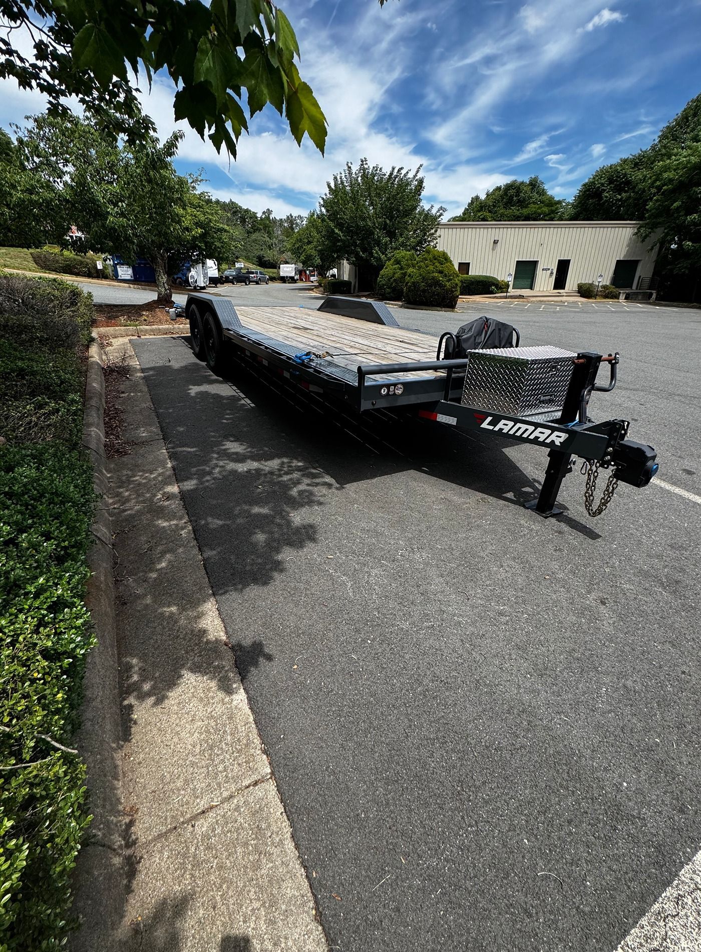 Black flatbed trailer parked on a suburban street beside a curb and trees under a blue sky.