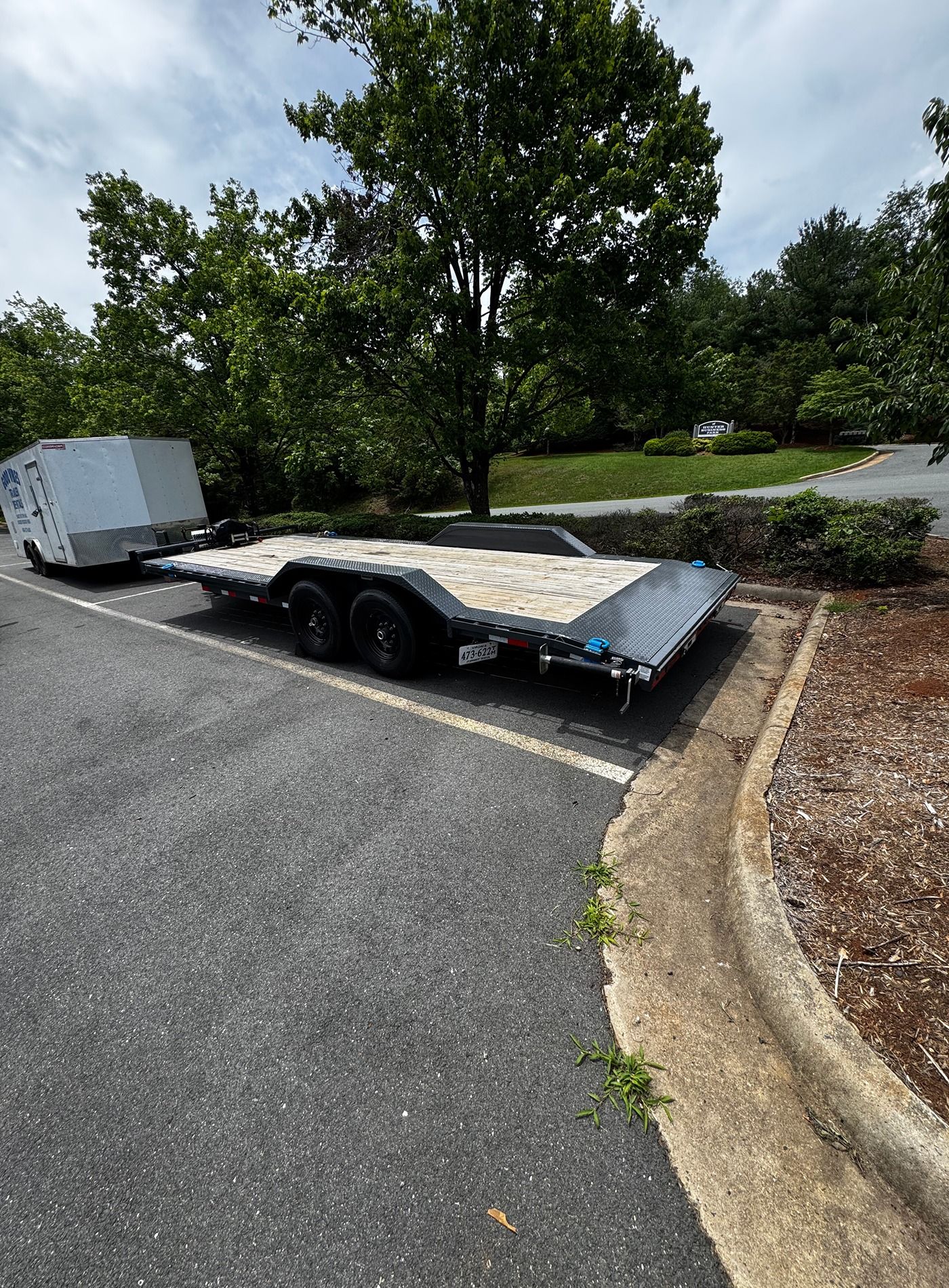 Flatbed trailer parked on asphalt beside a curb and trees, carrying a white enclosed trailer.
