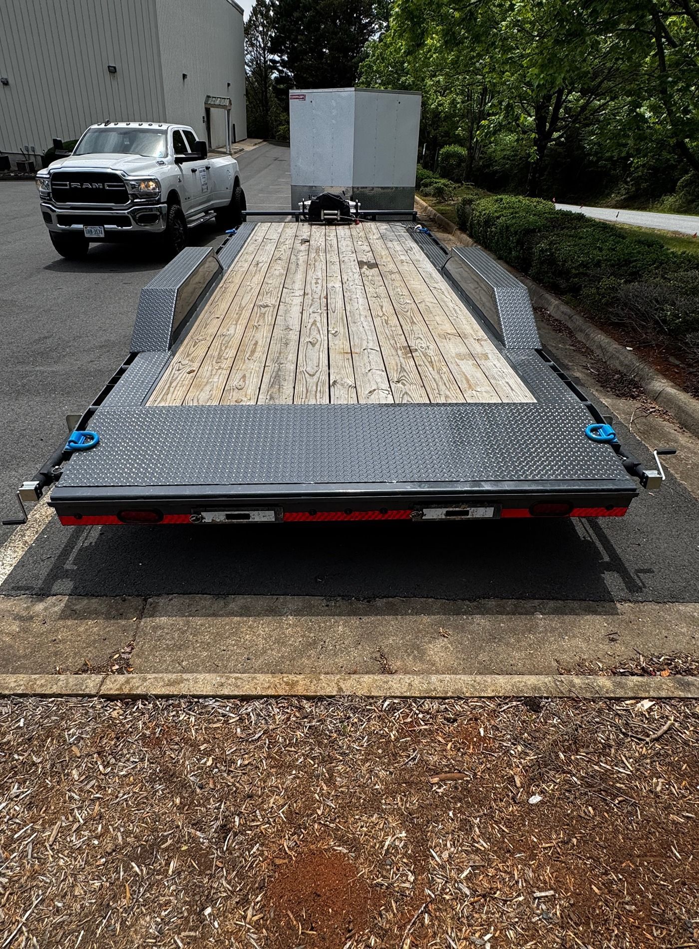 Flatbed trailer with wooden deck parked on a roadside, with a white truck and trees in the background.