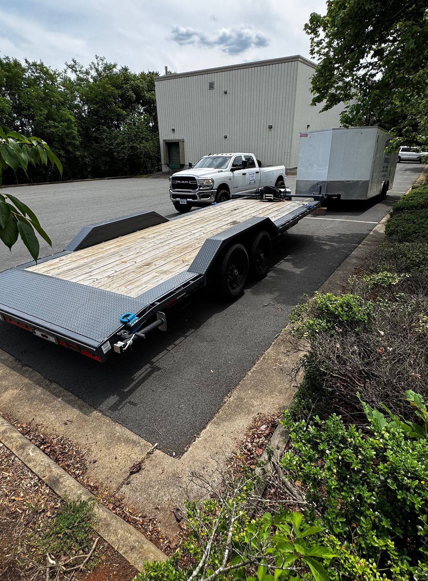 White pickup truck towing a flatbed trailer with a large white box on an asphalt lot near trees and a building