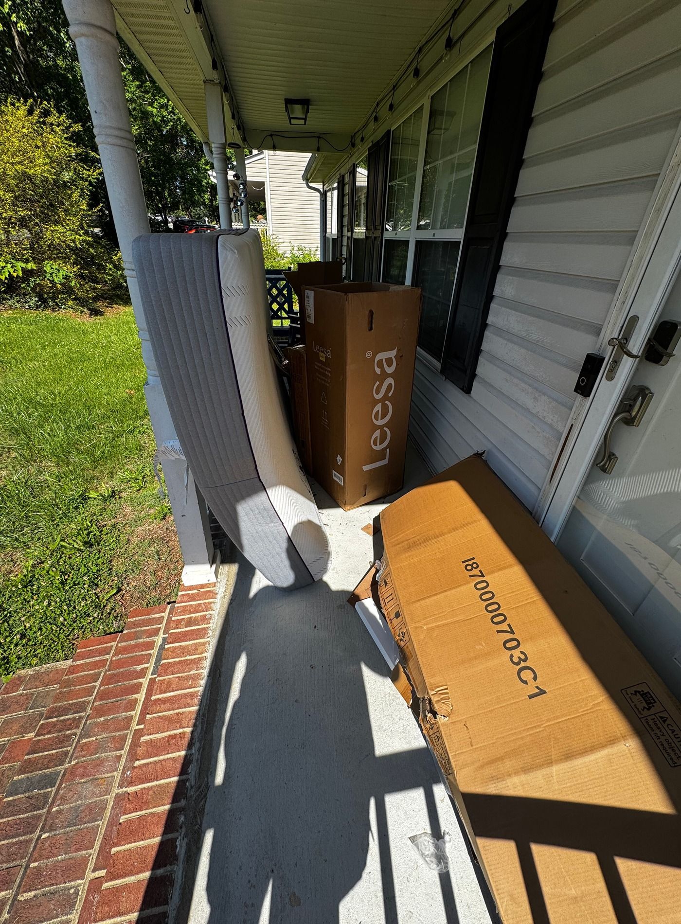 Packages on a front porch beside a white house and brick walkway, with a long shadow cast across the floor.