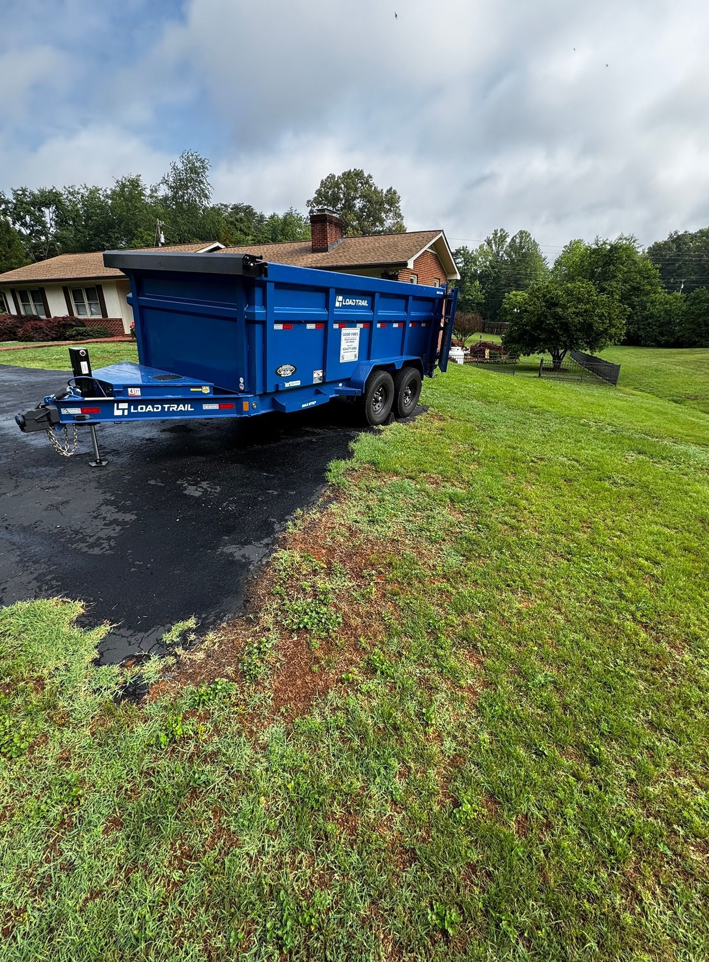 Blue dump trailer on a wet driveway beside a grassy yard and house under cloudy skies