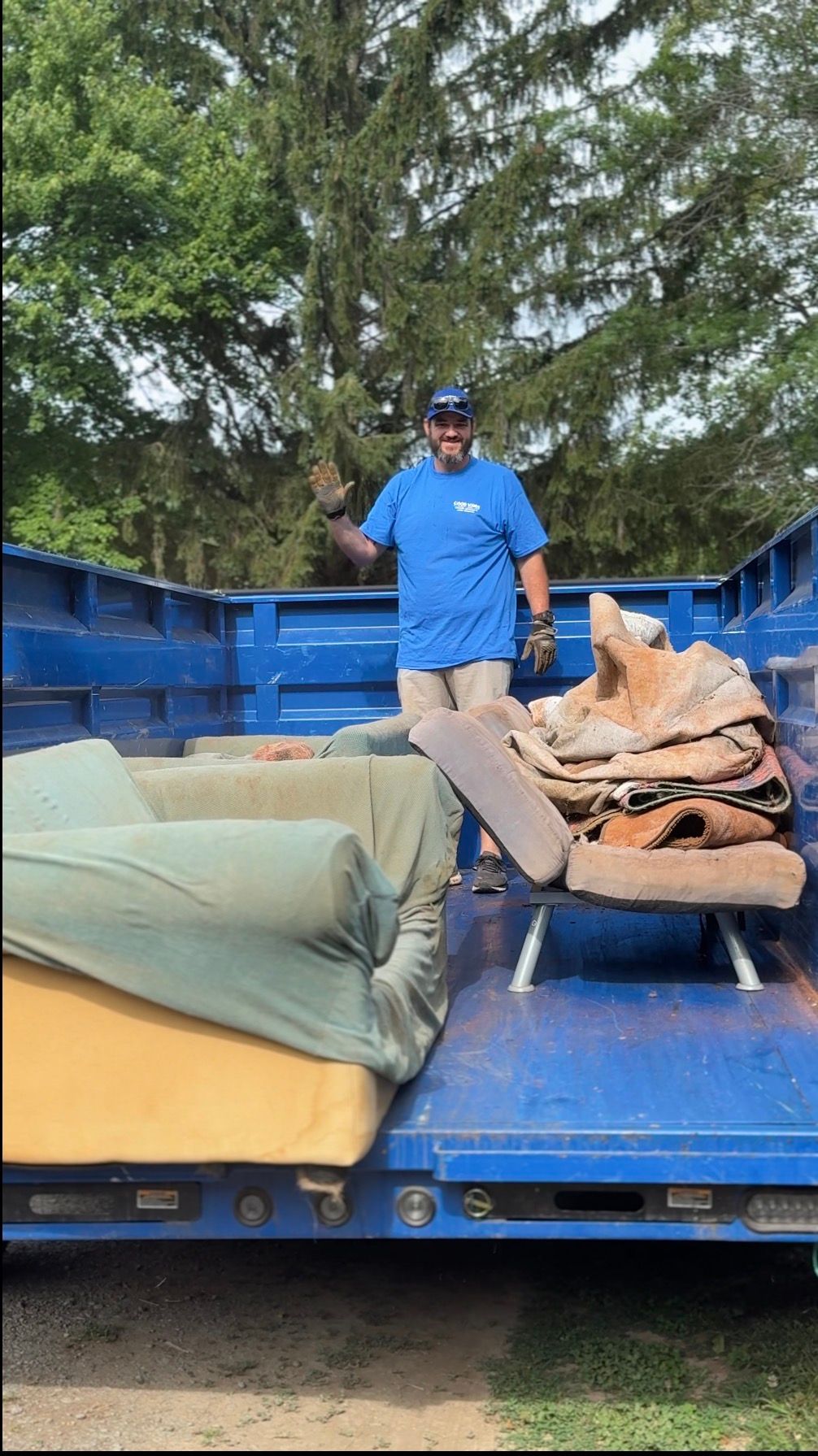 Man standing on a blue trailer loaded with bundled bags and a mattress outdoors under trees