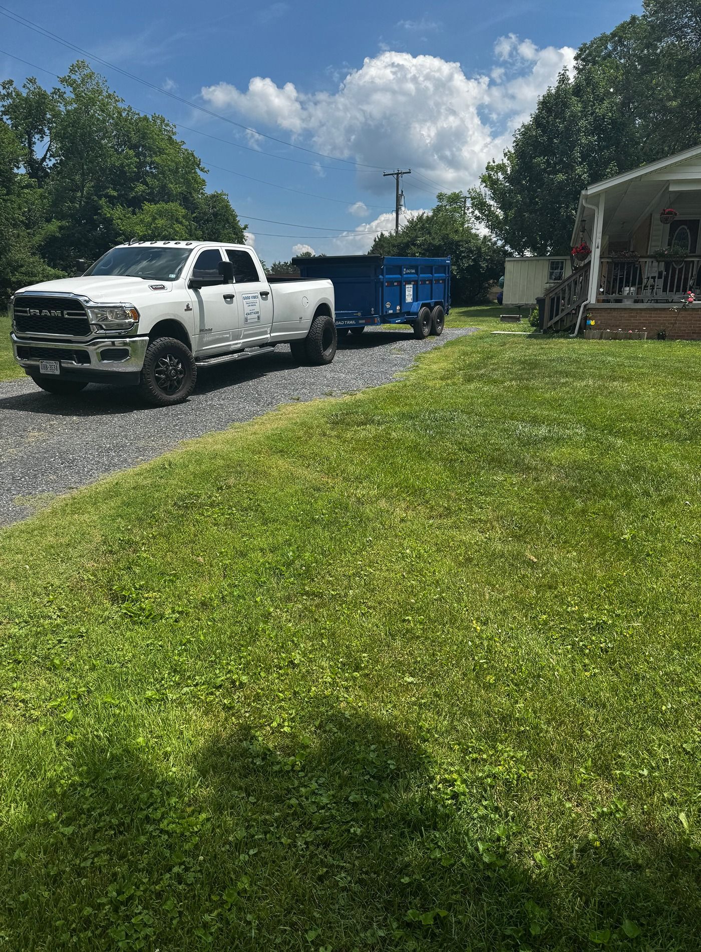 White pickup truck parked on a gravel driveway beside a blue dumpster and house under a sunny sky