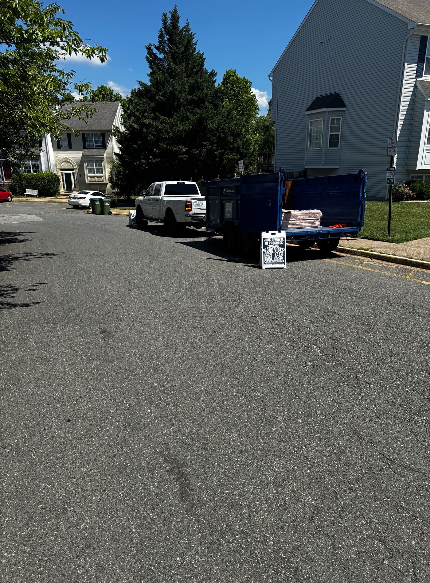 Residential street with parked white SUV and blue dumpster beside houses on a sunny day