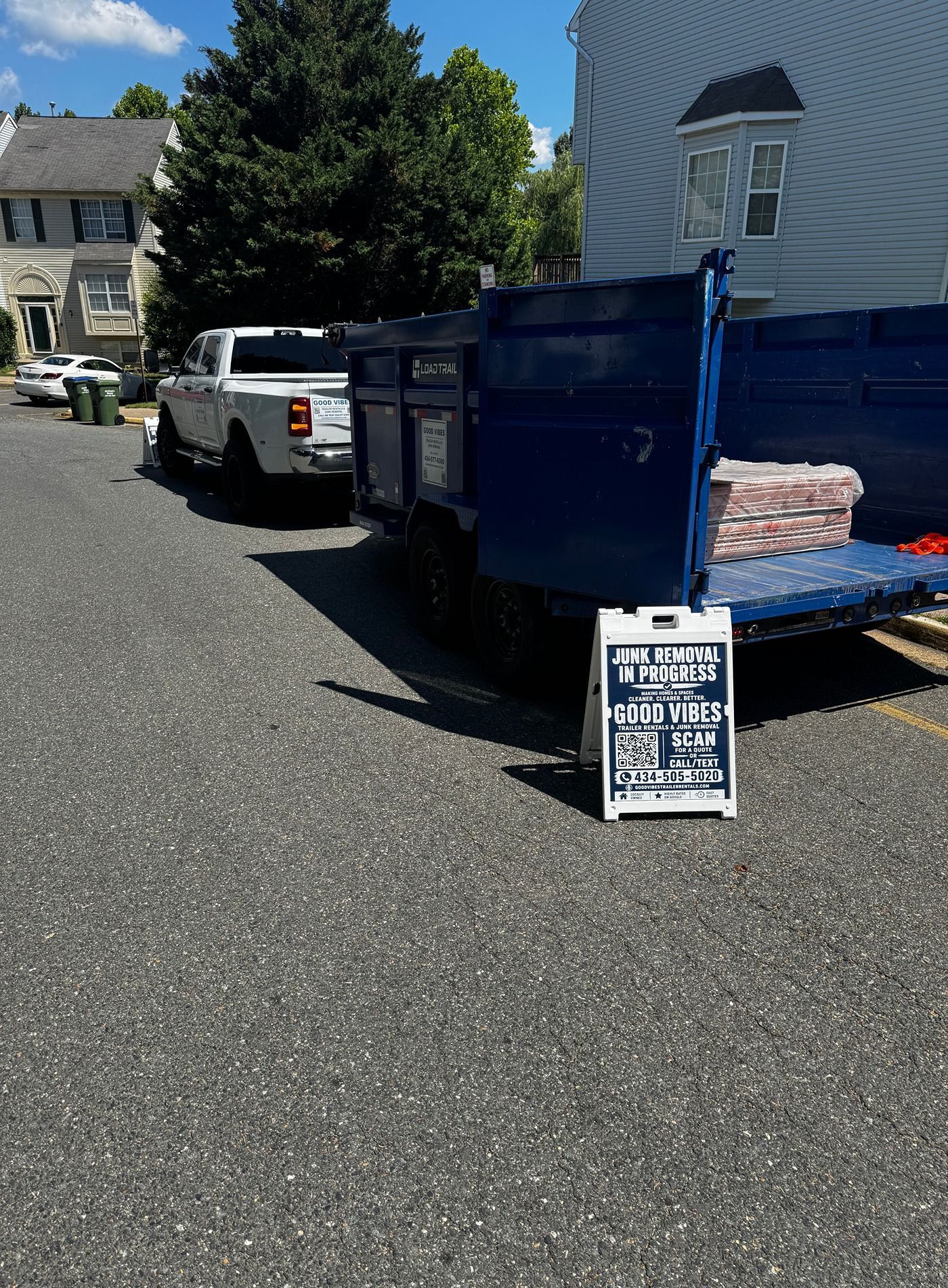 Blue dump trailer with a yard sign parked on a residential street beside a white truck.