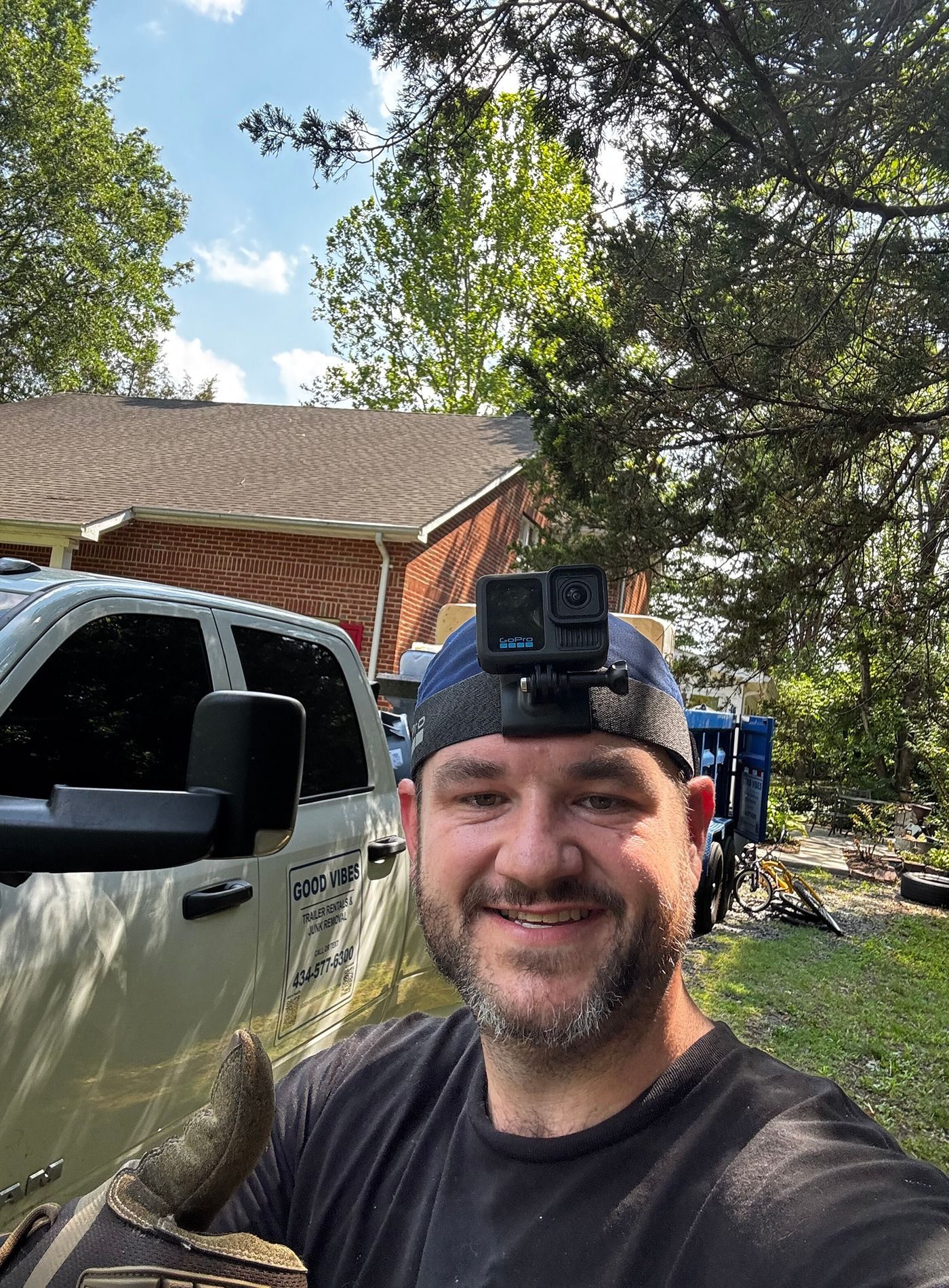 Smiling man wearing a head-mounted camera selfie beside a white truck and red barn in a wooded yard