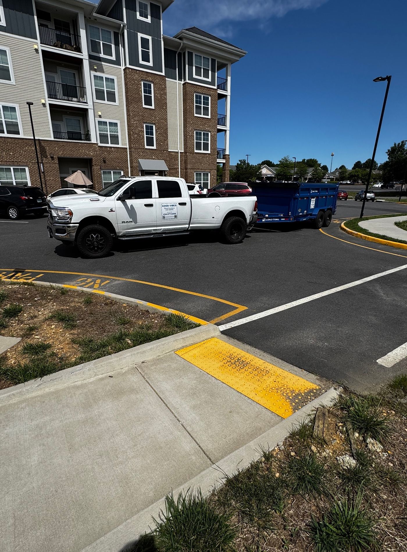 White pickup truck towing a blue trailer parked in a lot beside an apartment building and curb ramp