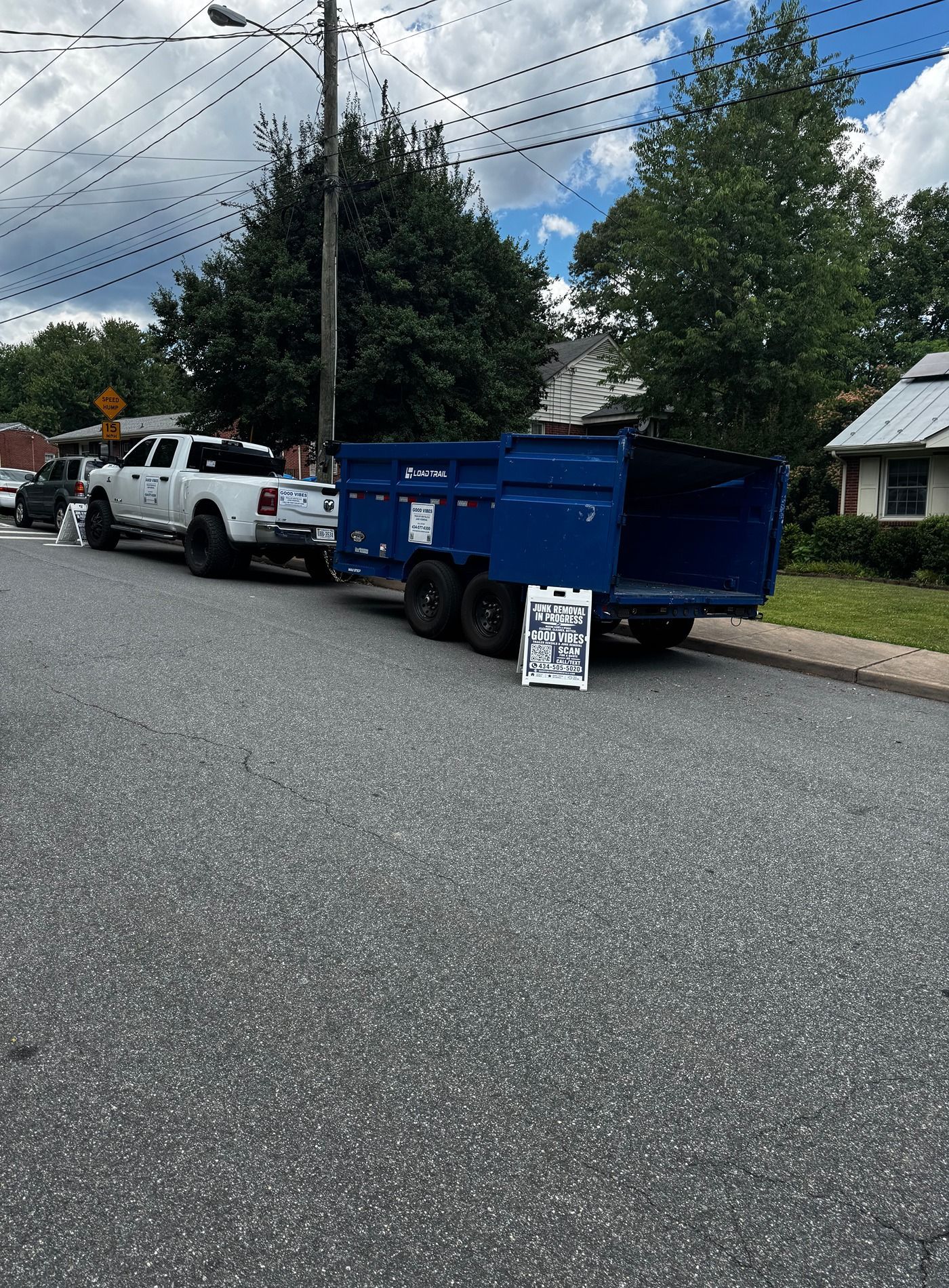 Blue utility trailer on a suburban street beside a white pickup truck and a small signboard.