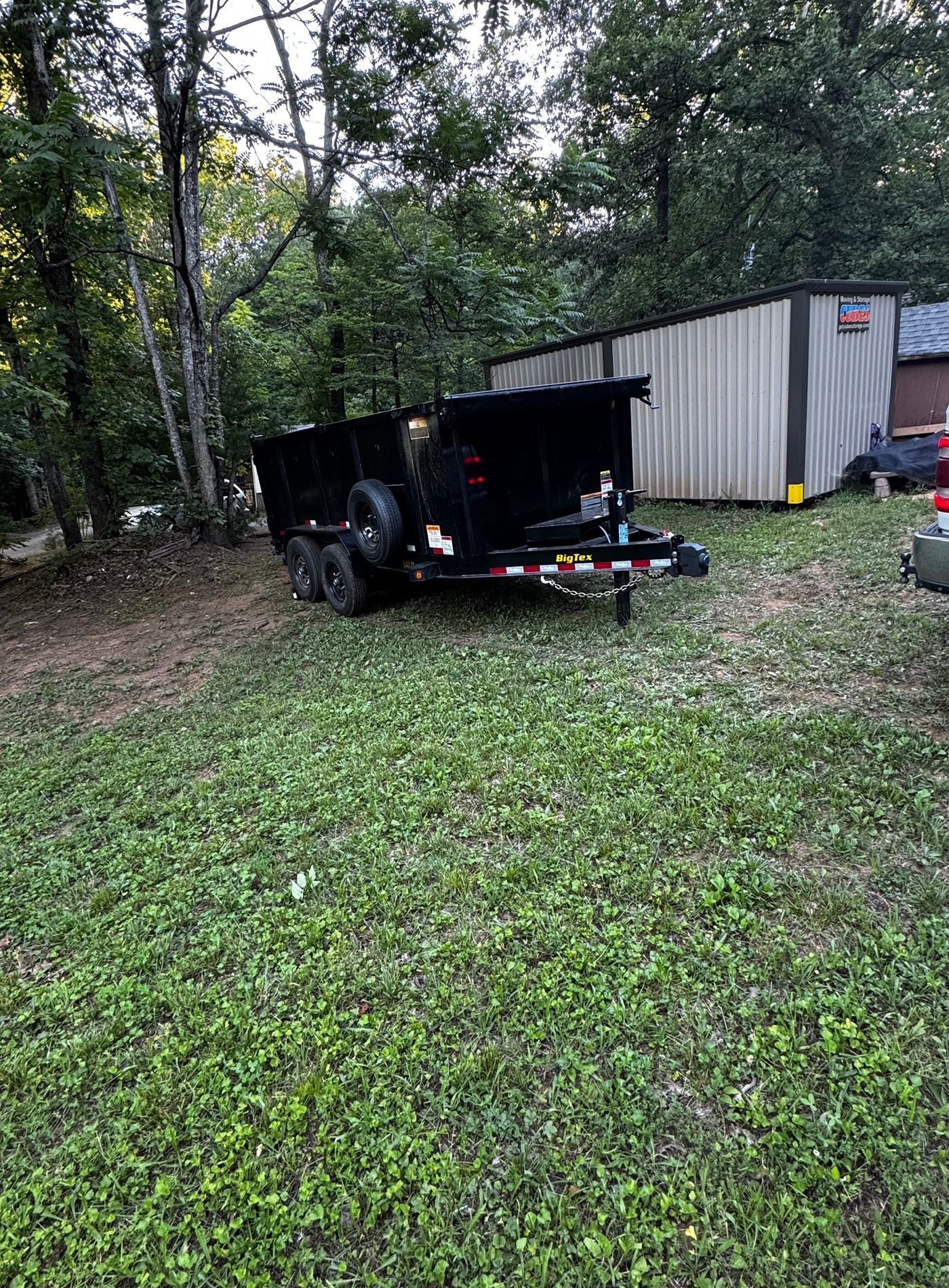 Black utility trailer parked on grass beside a white storage container in a wooded yard.