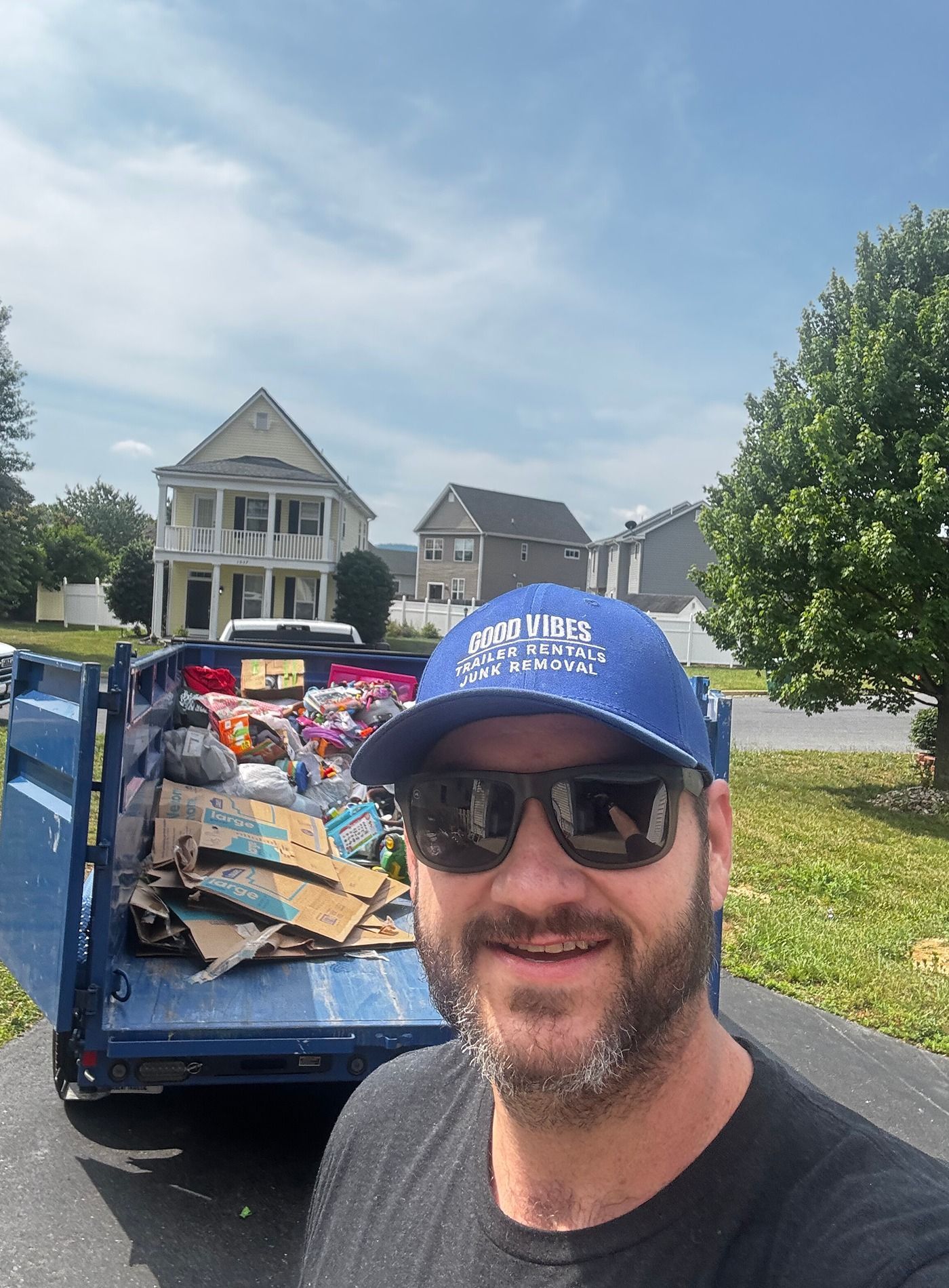 Man in blue cap taking a selfie beside a truck loaded with debris in a suburban neighborhood