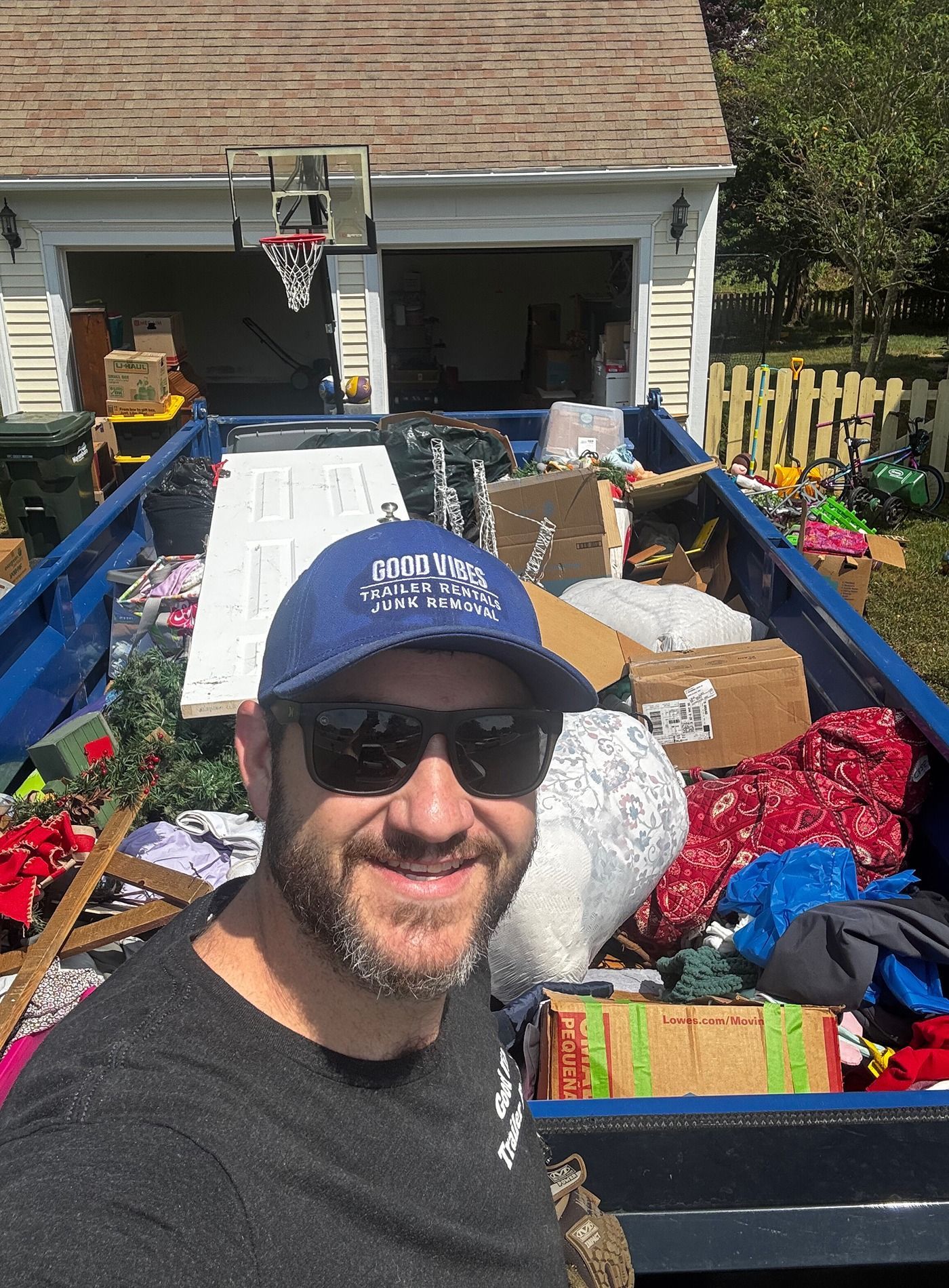 Smiling man in sunglasses and blue cap in front of a packed trailer in a driveway