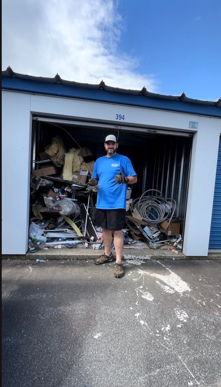 Man standing in front of a garage filled with clutter and debris, wearing a blue shirt and cap.