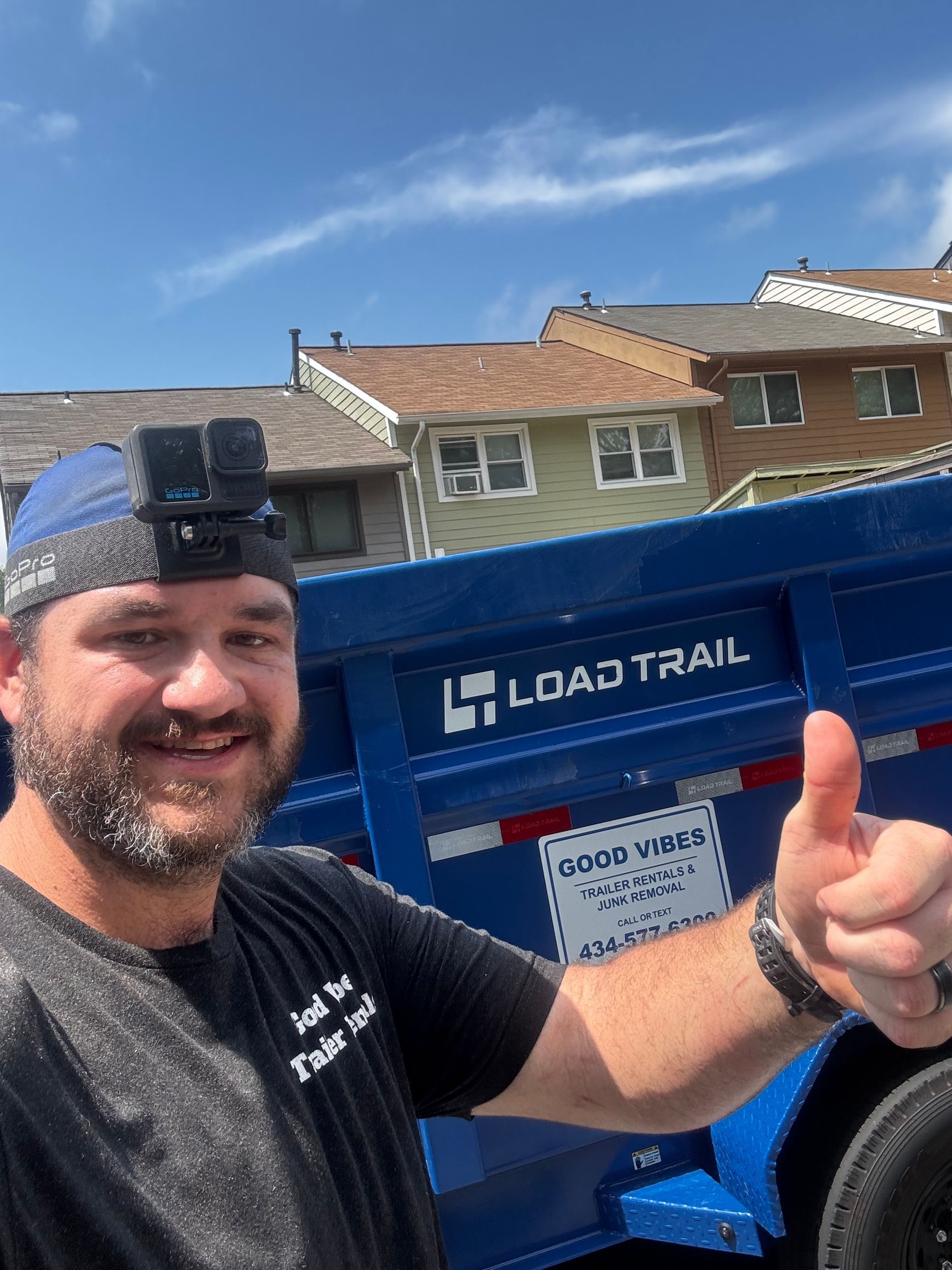 Smiling man in black shirt and blue cap giving thumbs up beside a blue Load Trail trailer.