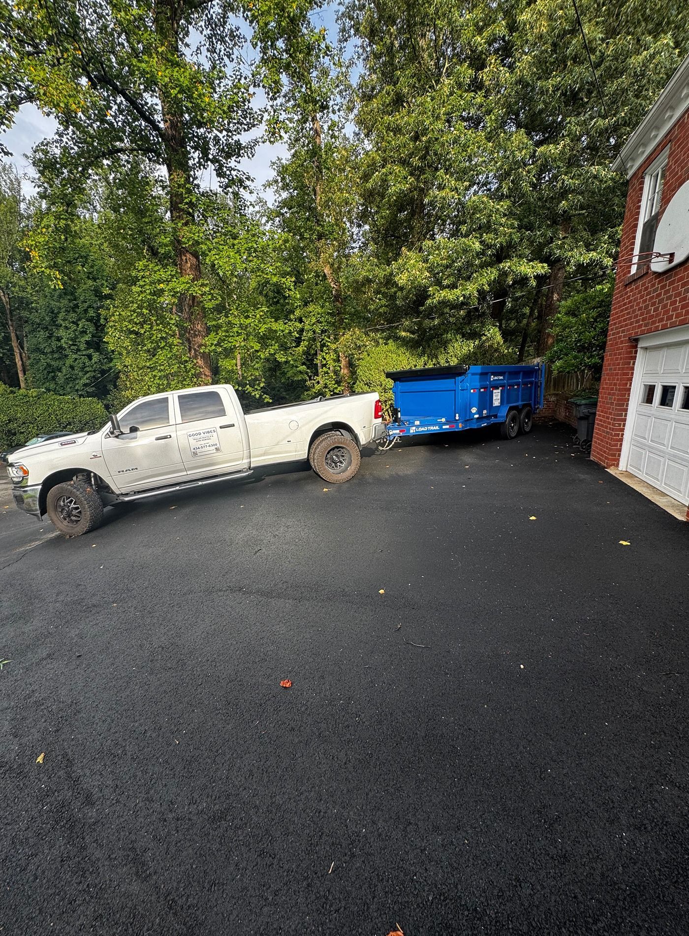 White pickup truck towing a blue trailer on a driveway beside a brick building and trees