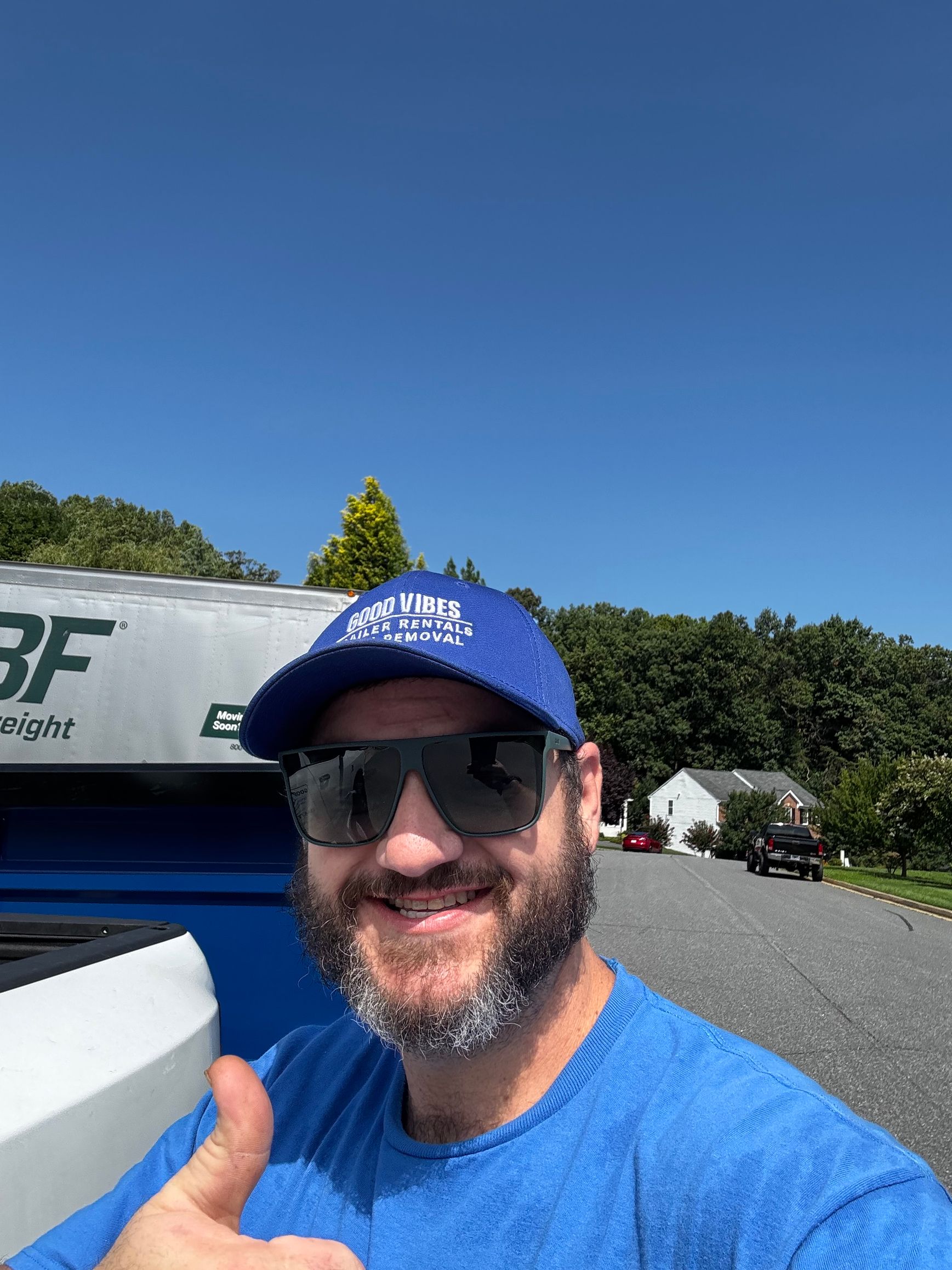 Man smiling in blue shirt and cap taking a selfie outdoors beside a building under a clear blue sky