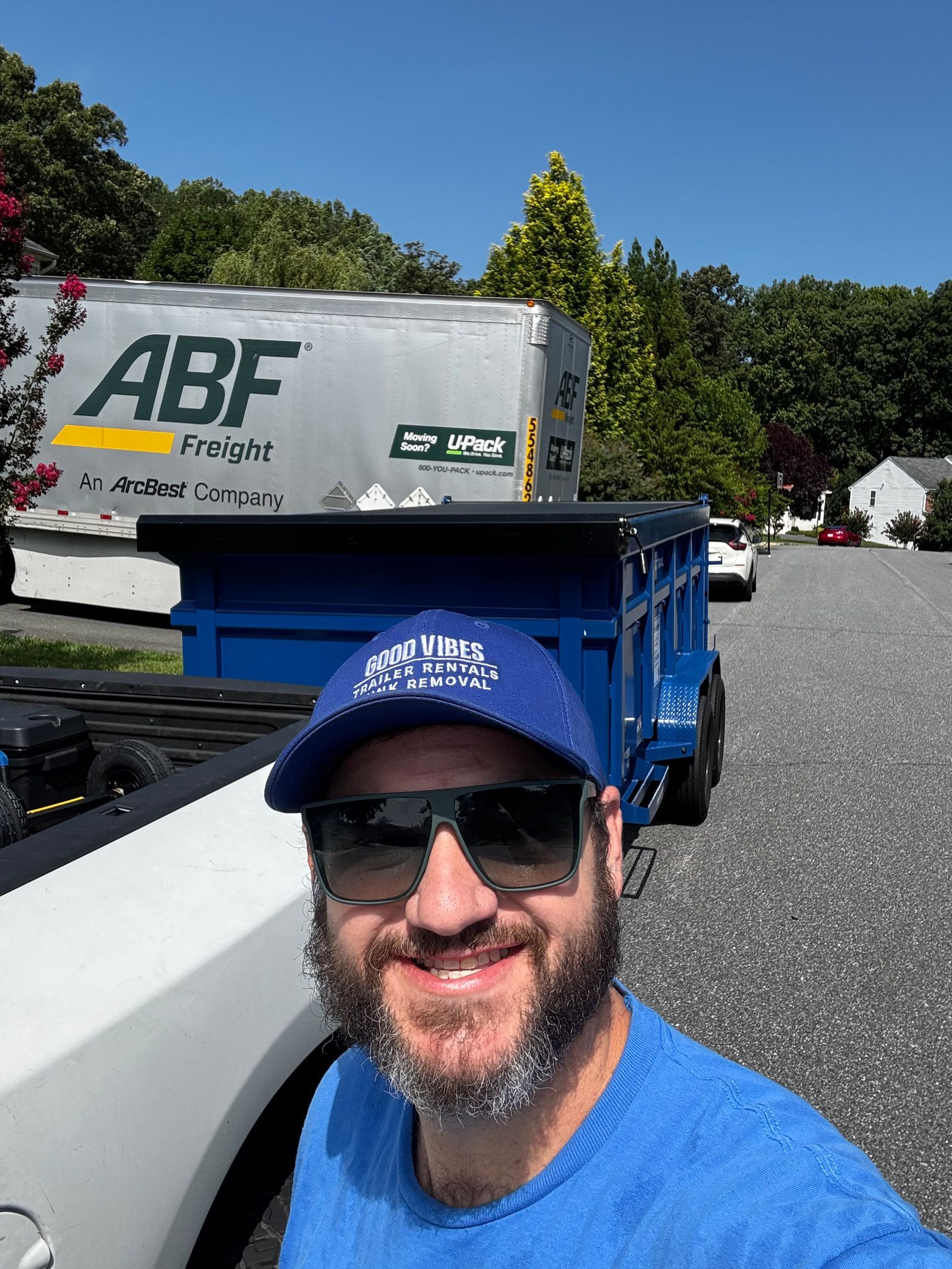 Man in blue cap and sunglasses taking a selfie beside a blue truck in a sunny parking lot
