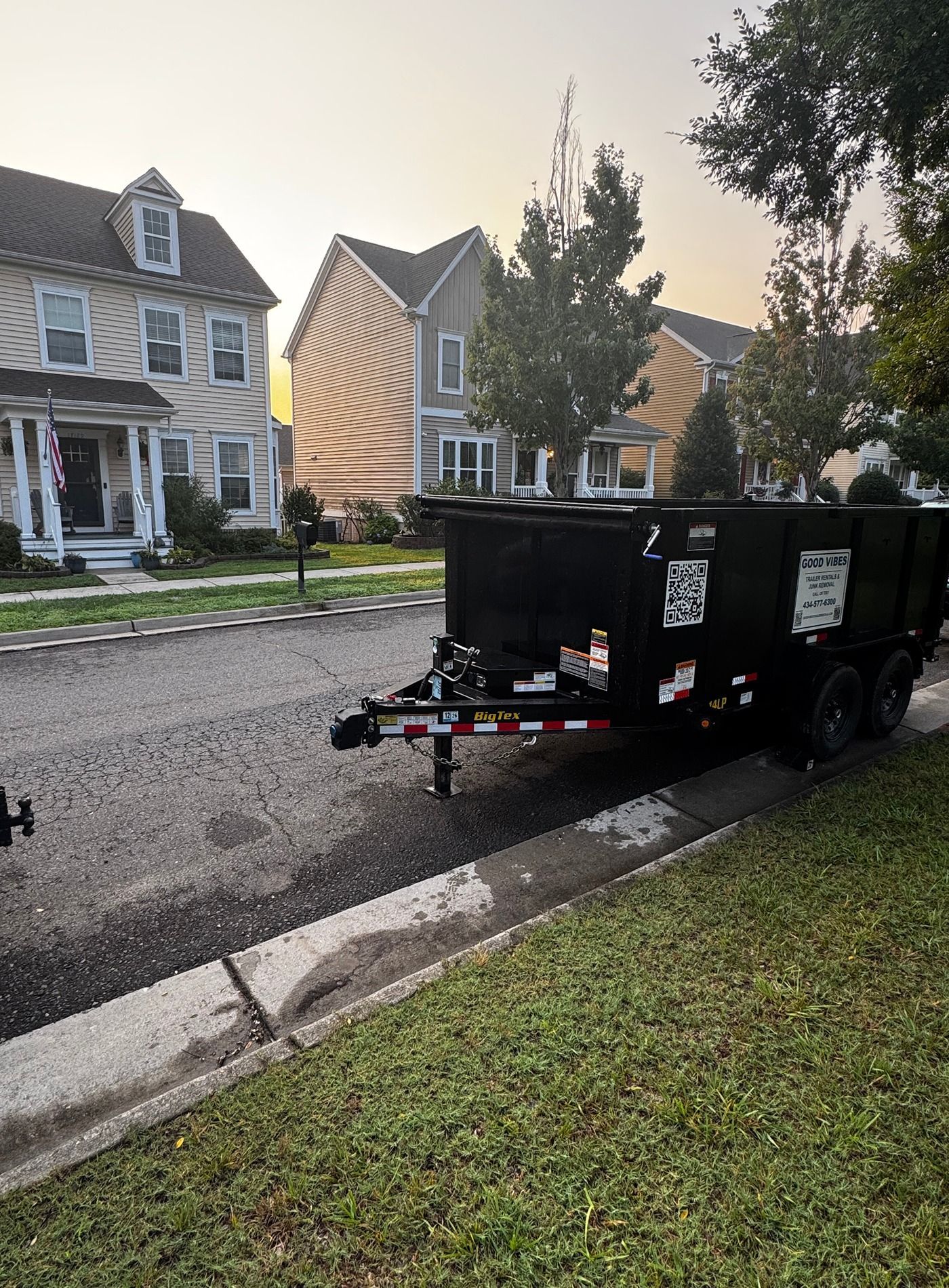 Black utility trailer parked by a curb on a residential street with houses and trees at dusk