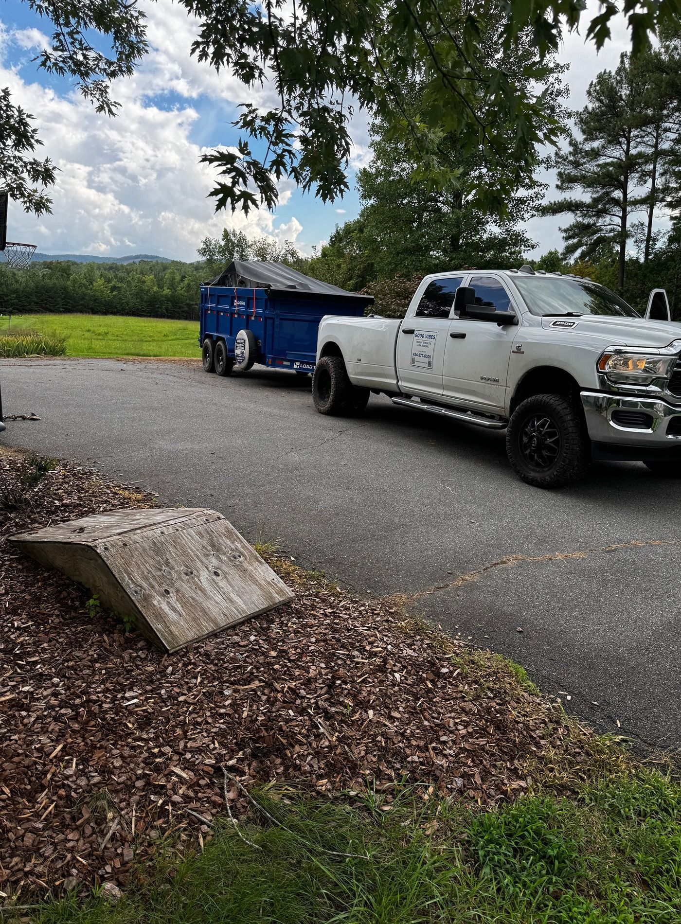 White pickup truck towing a blue trailer on a gravel driveway beside trees and a grassy field.