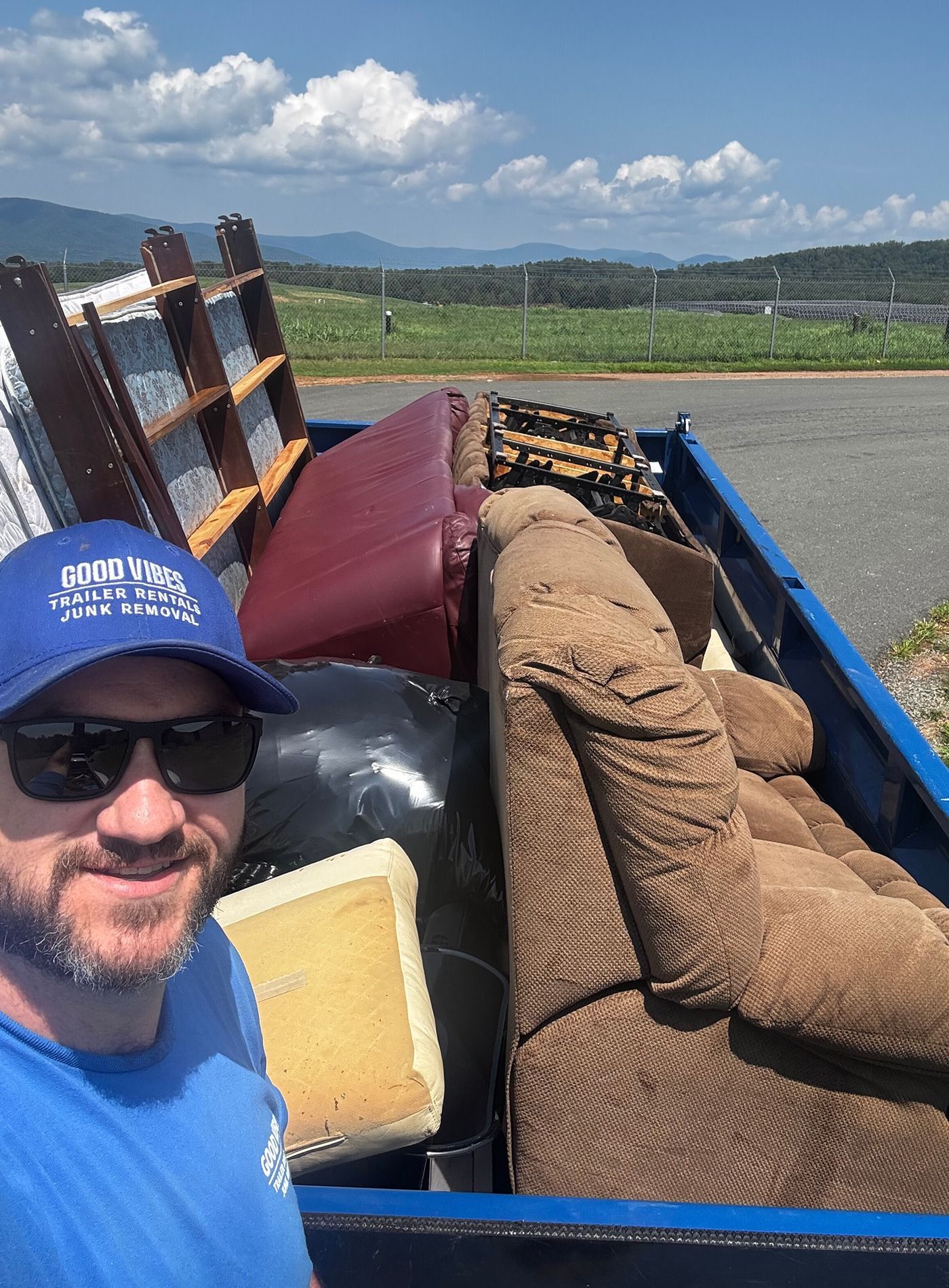 Man in blue shirt and cap taking a selfie beside a truck loaded with furniture in a rural field