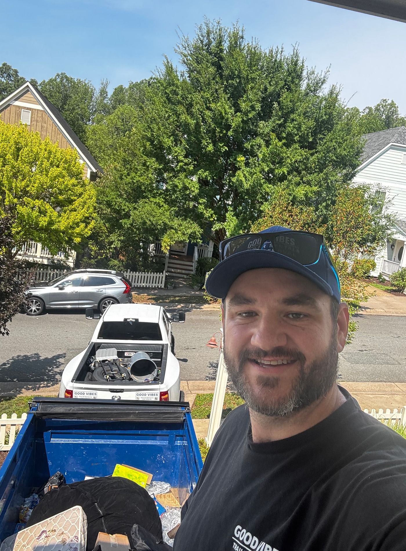 Man smiling in black cap and shirt on a truck bed, with a white car and trees in a sunny suburban street.
