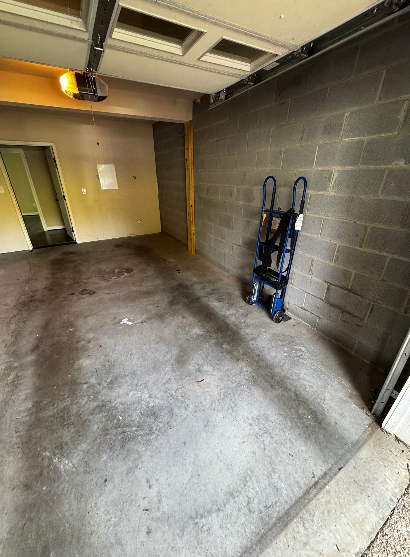 Empty garage with concrete floor, cinderblock wall, and a blue hand truck leaning against the wall.