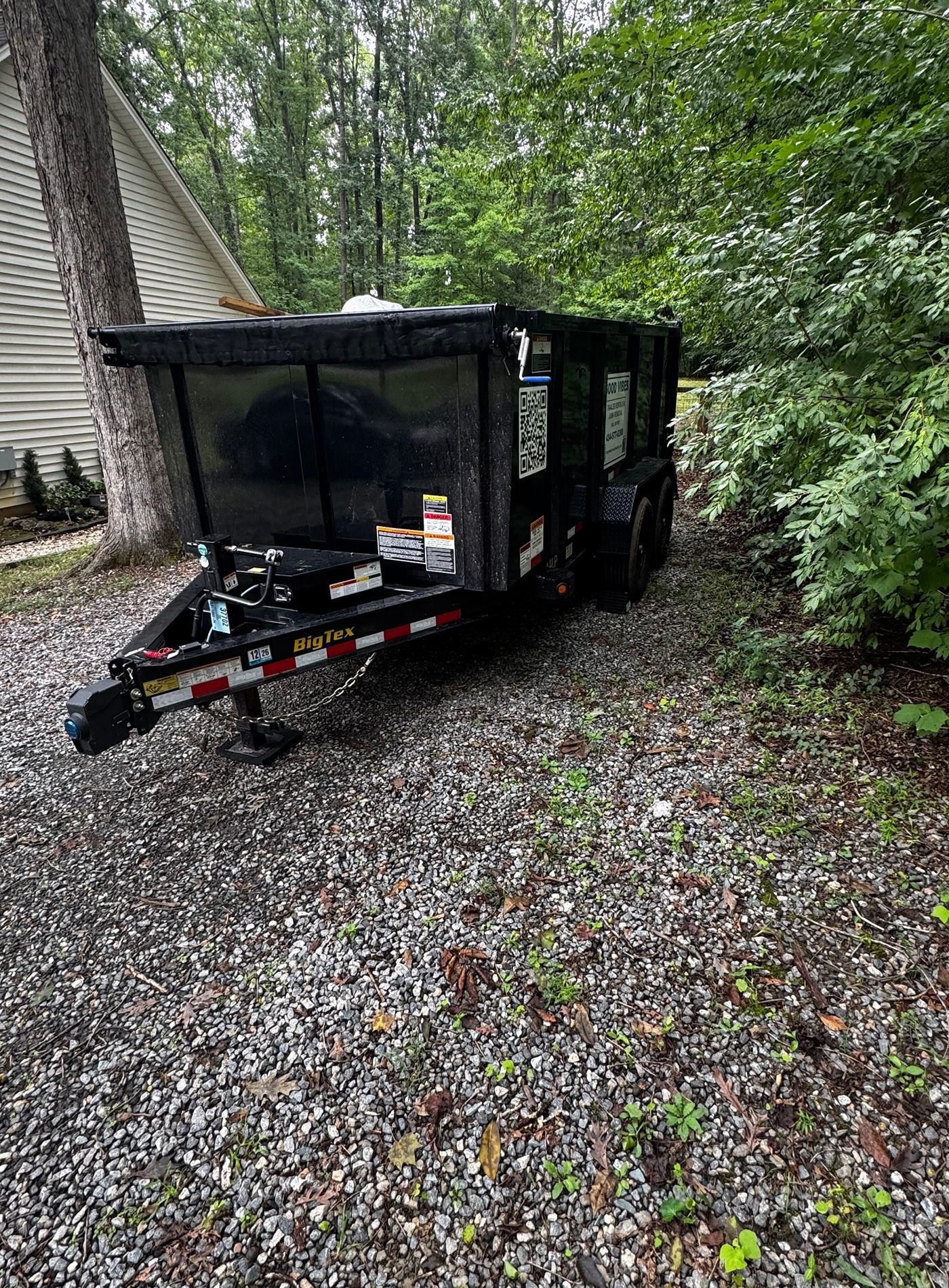 Black enclosed utility trailer parked on gravel beside a house in a wooded yard