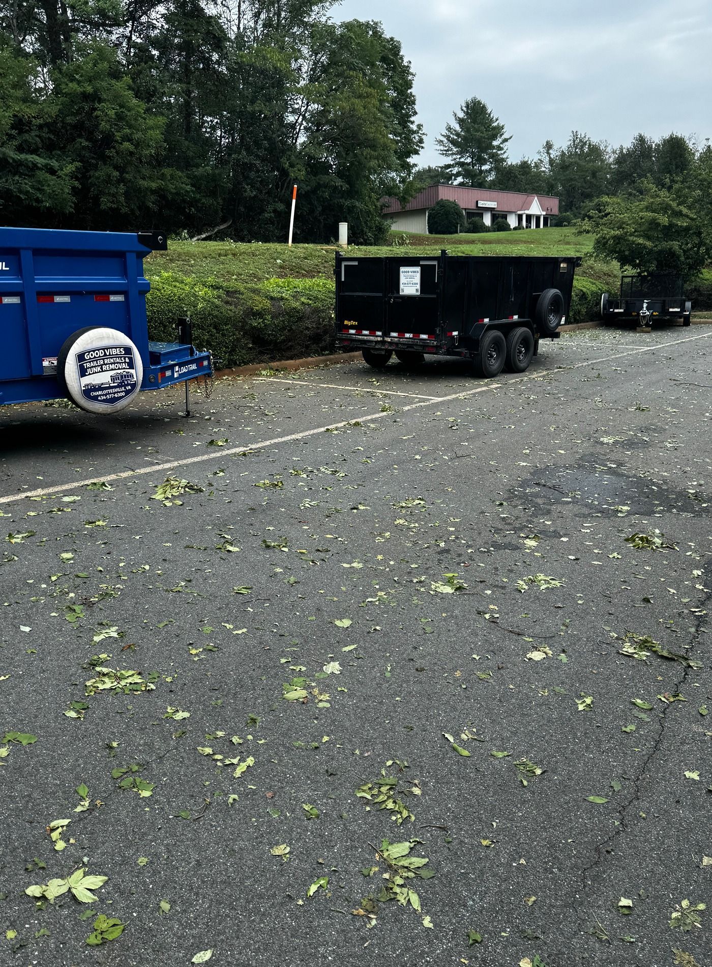 Blue and black utility trailers on a gravel lot with scattered leaves, trees and buildings in the background