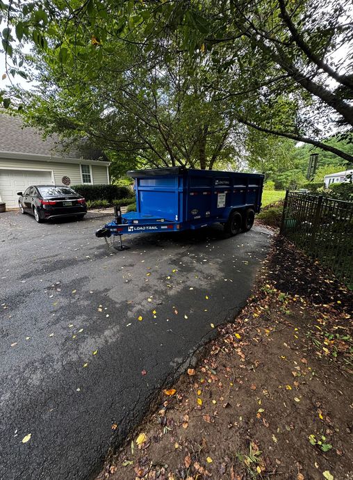 Blue utility trailer parked on a wet residential driveway under leafy trees, with a black car nearby.