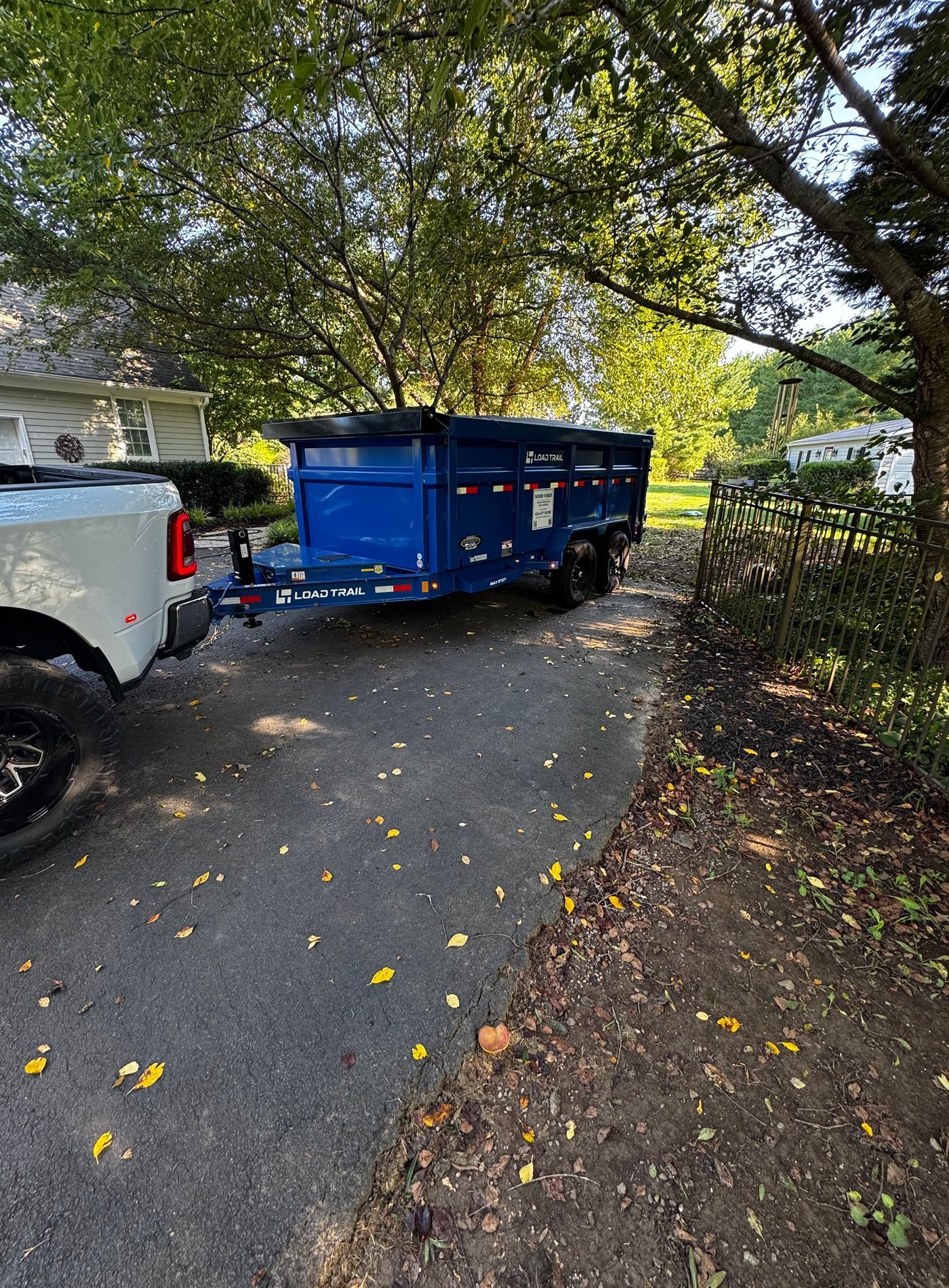 Blue dumpster trailer hitched to a white truck on a tree-lined driveway.