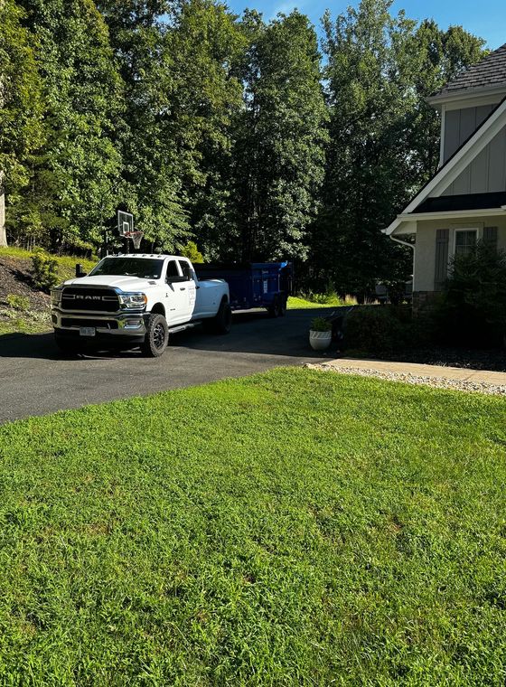 White pickup truck parked by a blue dumpster beside a house with a green lawn and trees.