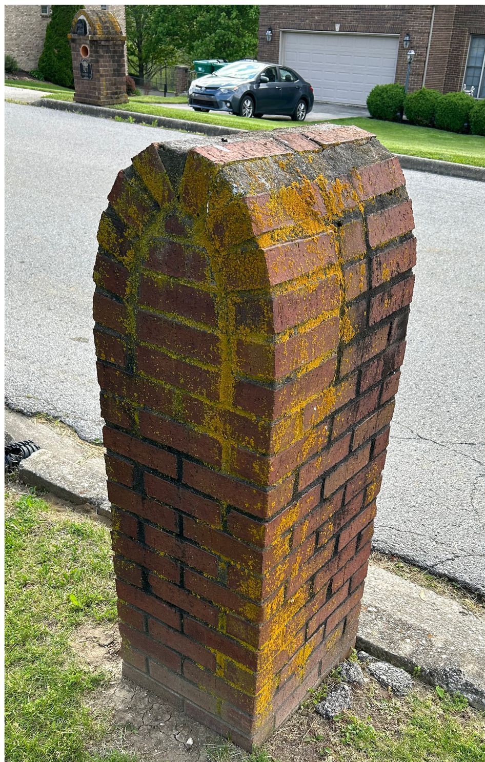 A brick post with moss on it is sitting on the side of the road.