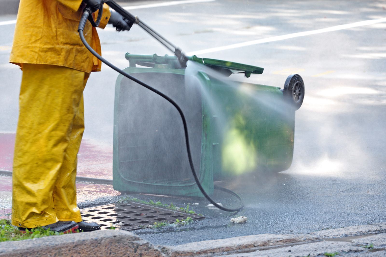 A man in yellow pants is cleaning a green trash can with a high pressure washer.