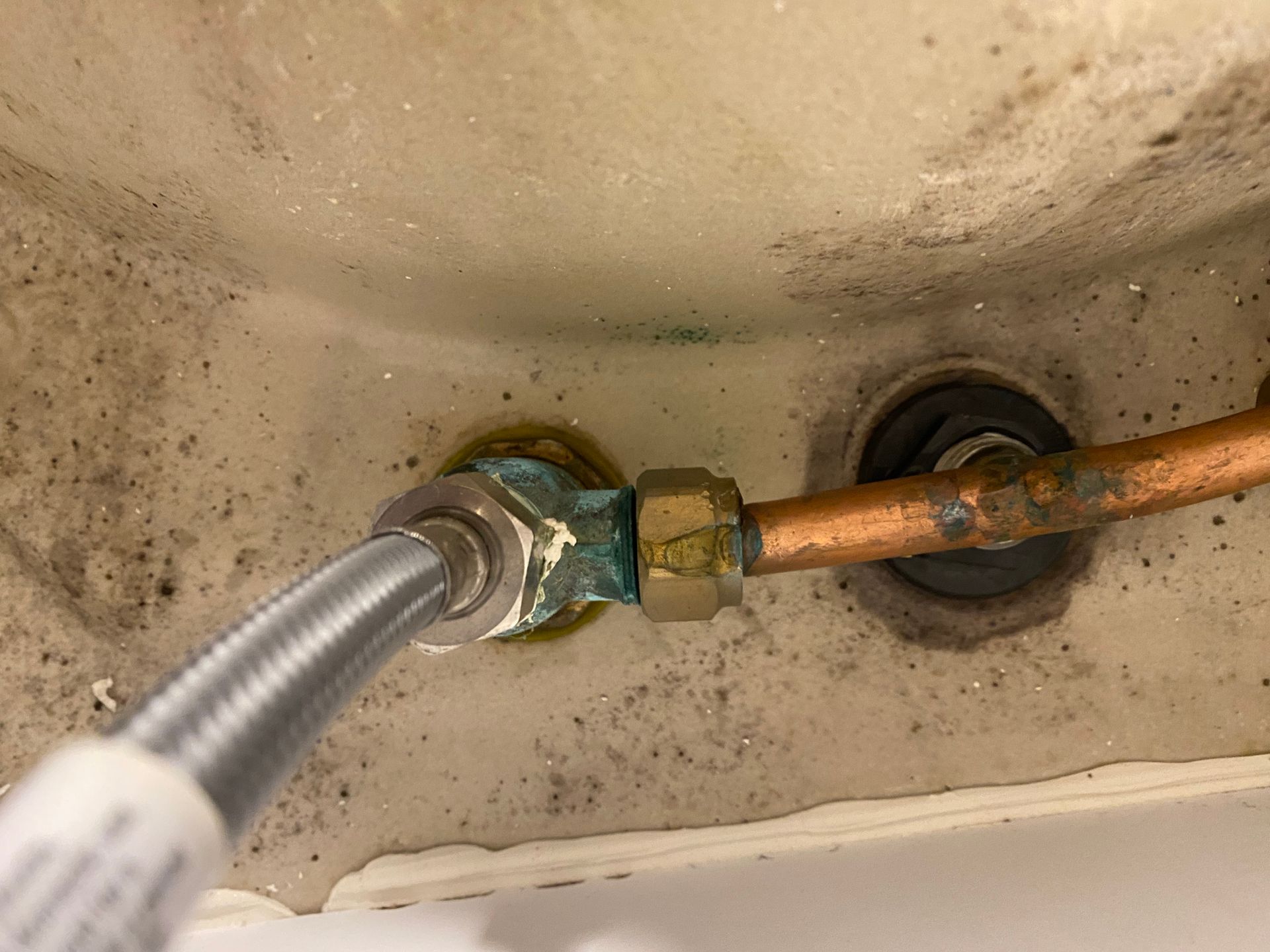 Close-up of copper plumbing connections under a sink; green corrosion on fittings, gray braided hose attached.